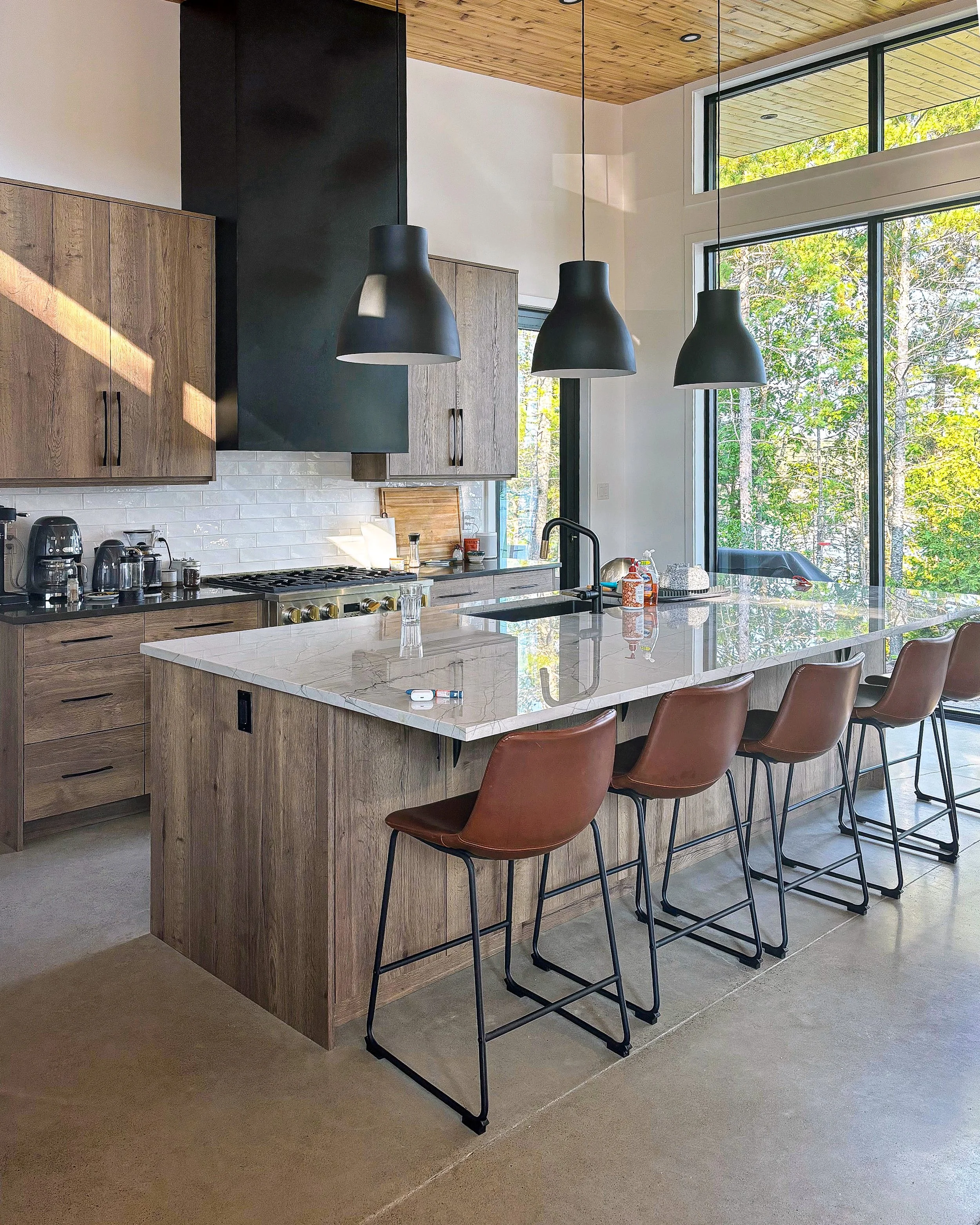 Modern cottage kitchen in the Kawarthas with wooden cabinets, steel hood vent, marble island, black pendant lights, large windows with a view of green trees, and various kitchen appliances.