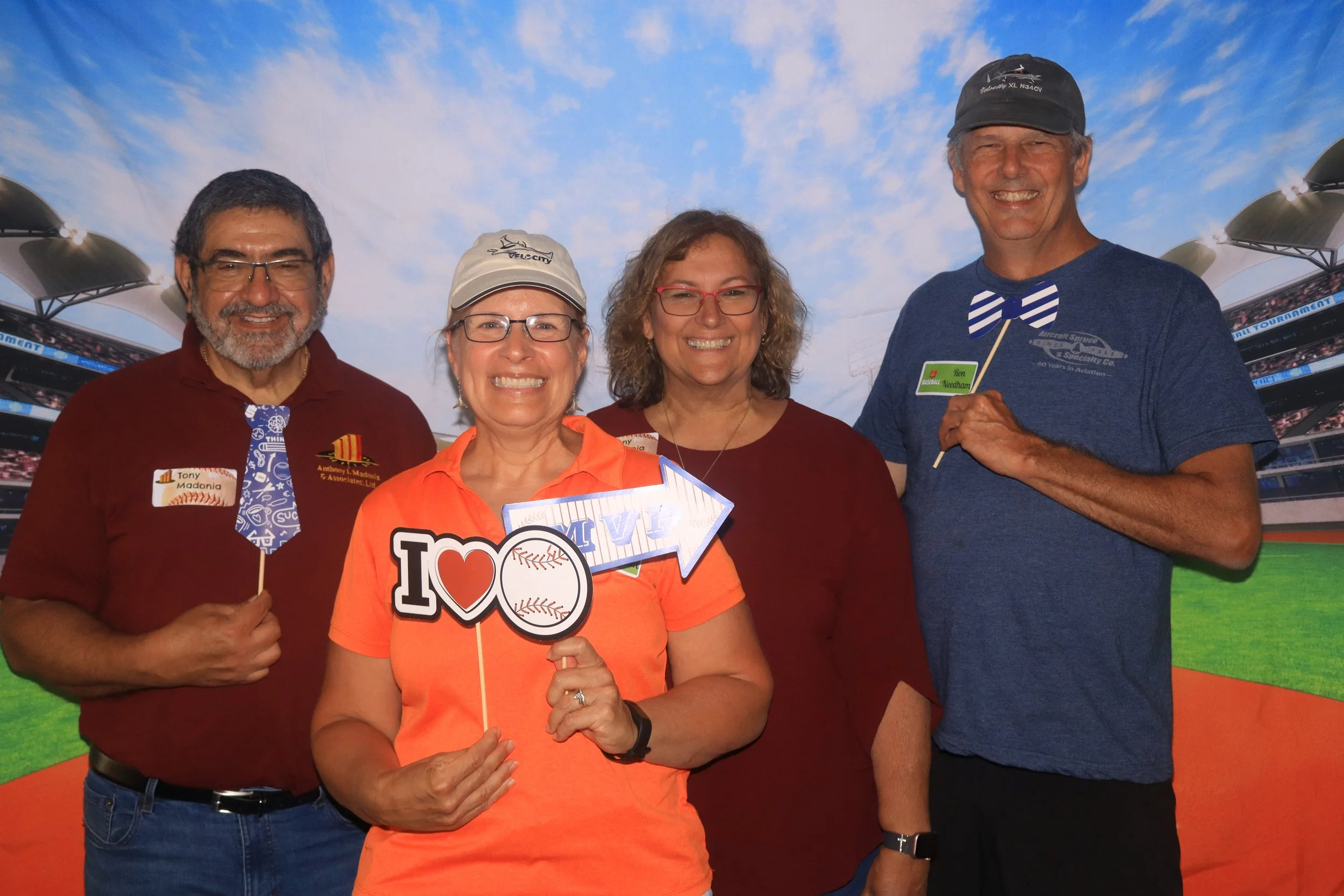 Four people standing together at a baseball stadium, smiling and holding signs, with a bright sky overhead.