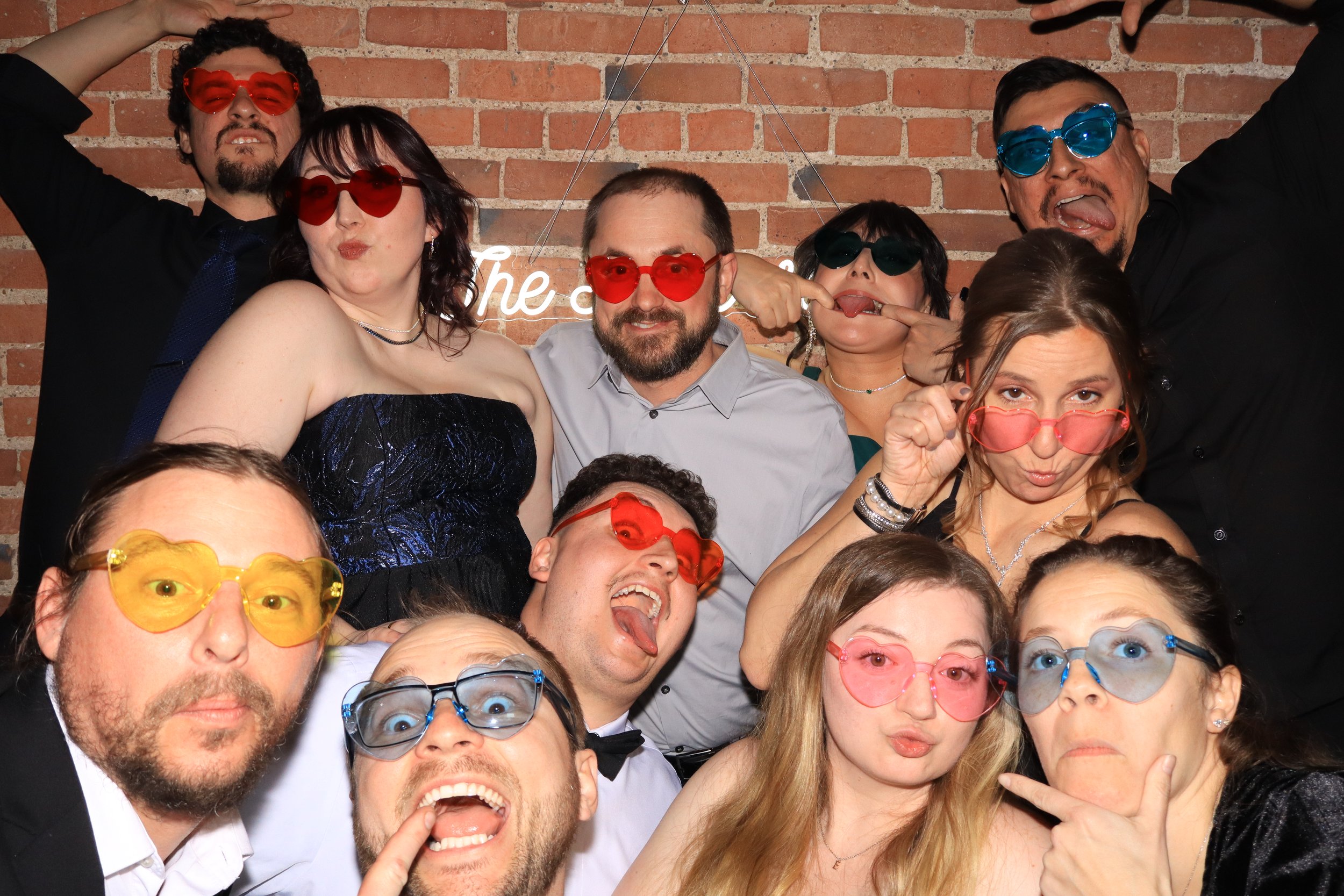 A group of diverse people wearing colorful sunglasses, smiling, making funny faces, and having fun against a brick wall background.