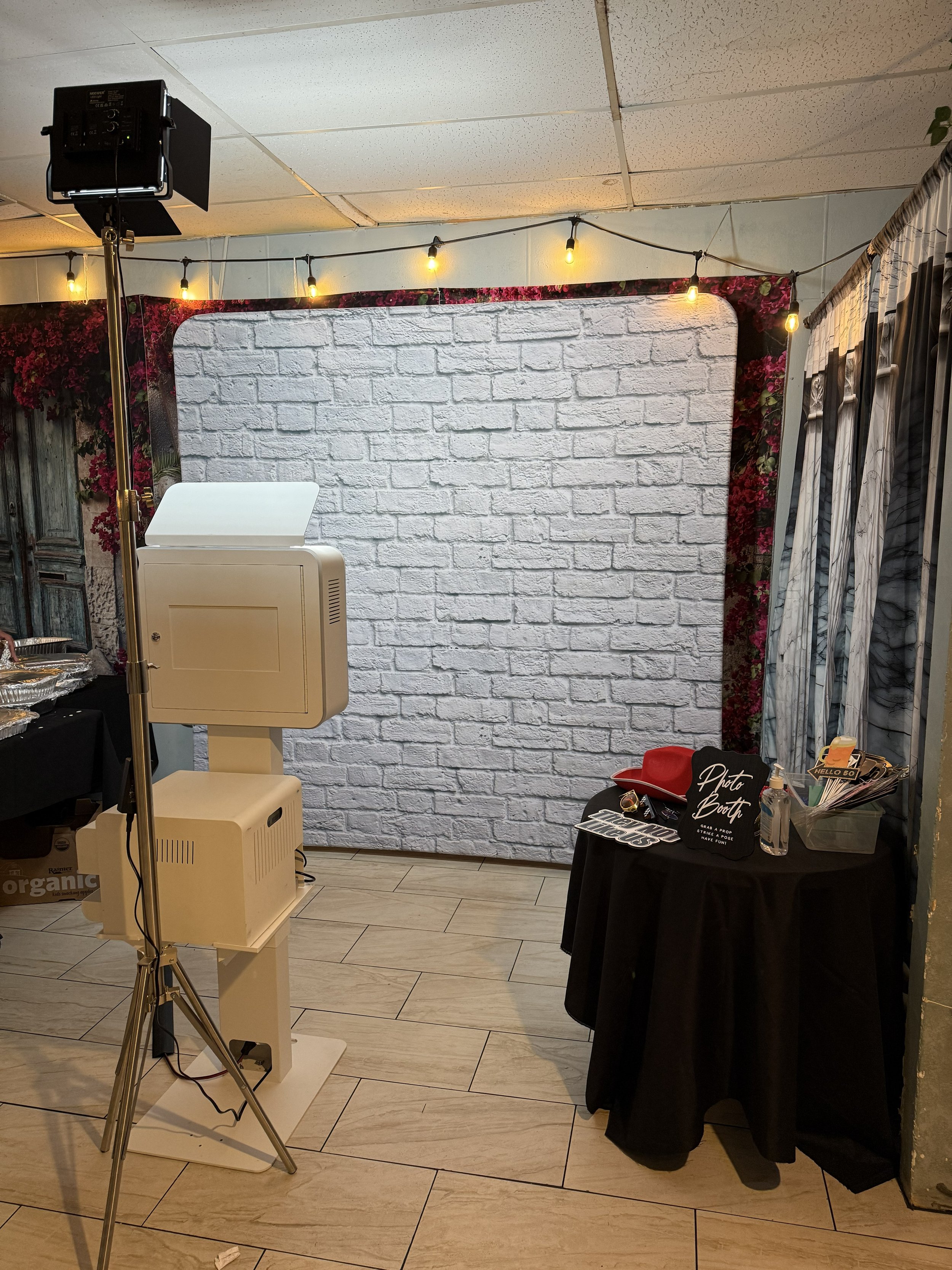 A photo booth setup with a white brick backdrop, string lights overhead, and a small black round table with props like glasses, hats, and signs. A photo booth machine and a lighting stand are also visible.