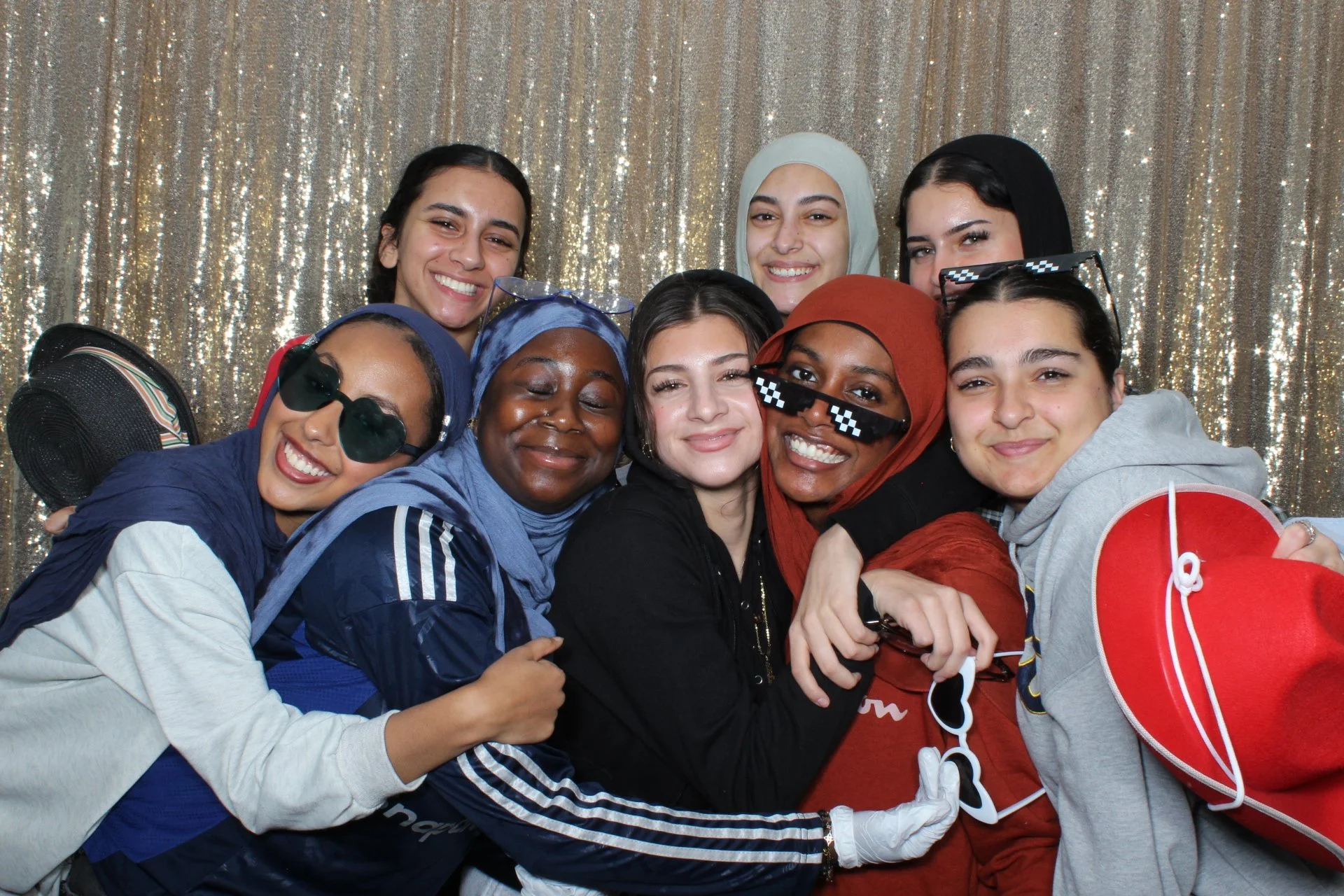 Group of women smiling and hugging in front of a sparkly gold curtain, wearing casual clothes, some with sunglasses, some with headscarves, holding hearts and a prop that looks like a paddle.