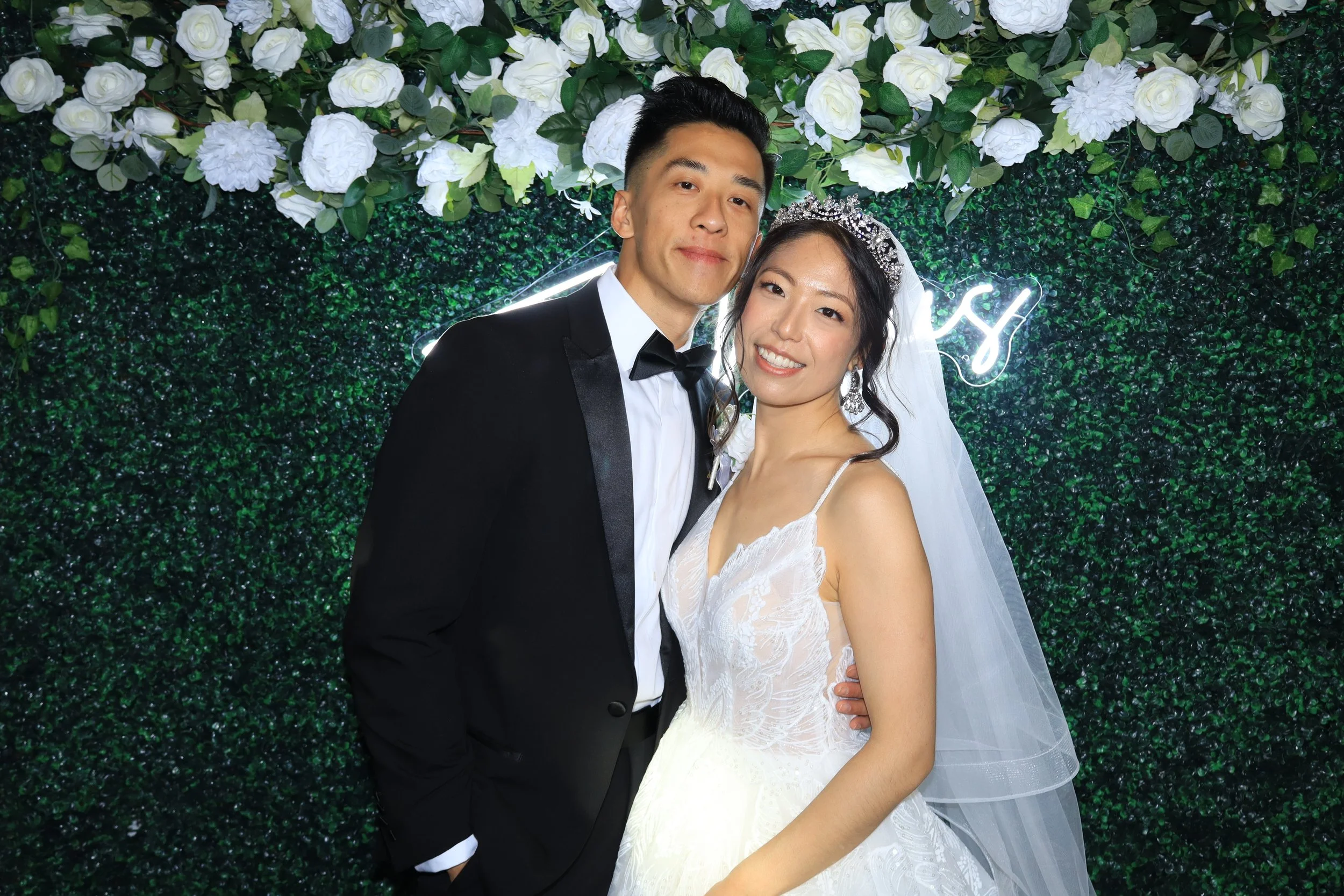 A wedding couple, a groom in a tuxedo and a bride in a wedding dress with a veil and tiara, smiling and posing together in front of a decorated floral backdrop.