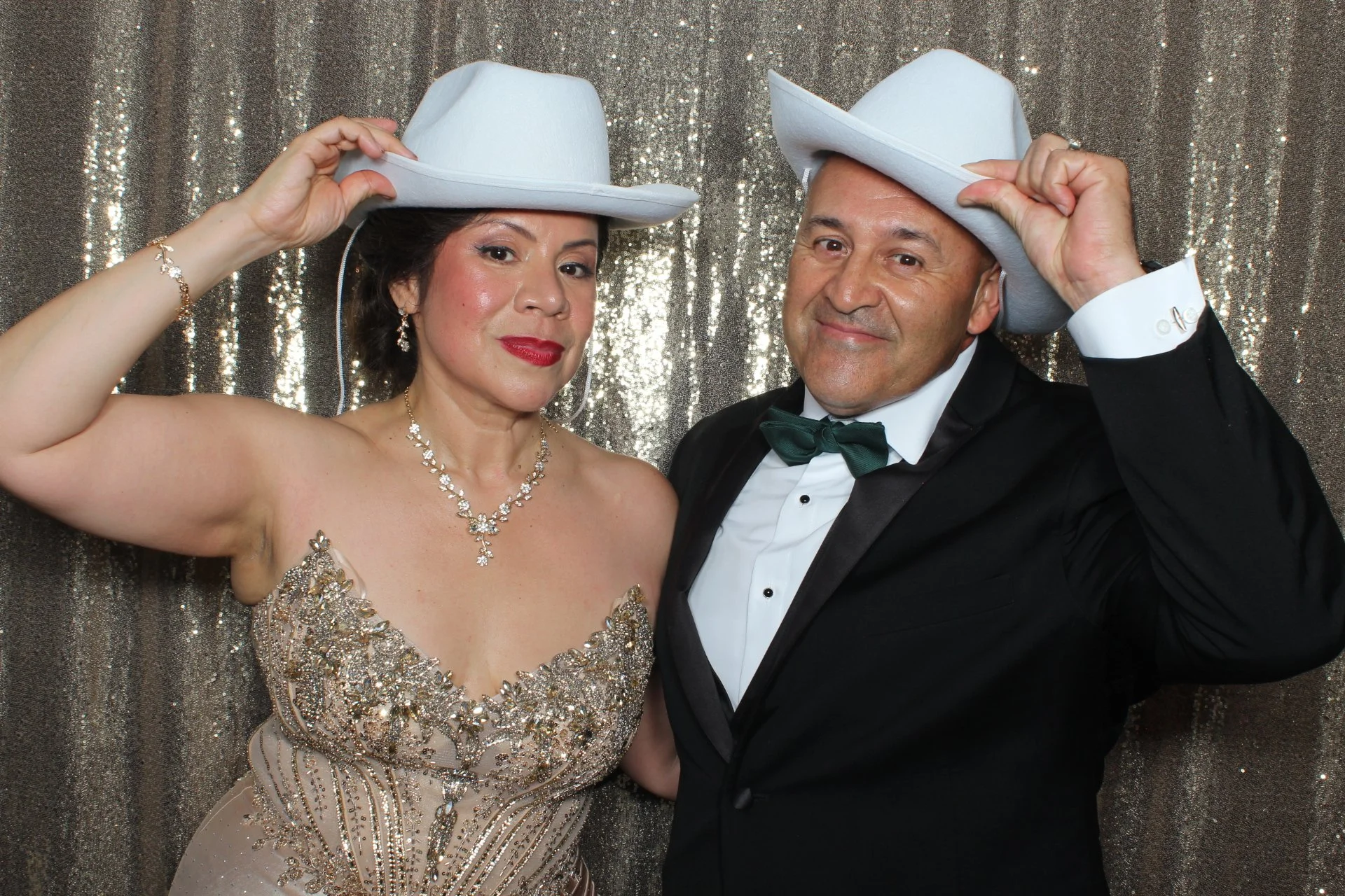 A couple in formal attire at a photo booth, wearing white cowboy hats, with glittery silver backdrop. The woman is in a glittering gold gown and the man is in a tuxedo with a bow tie, both smiling.
