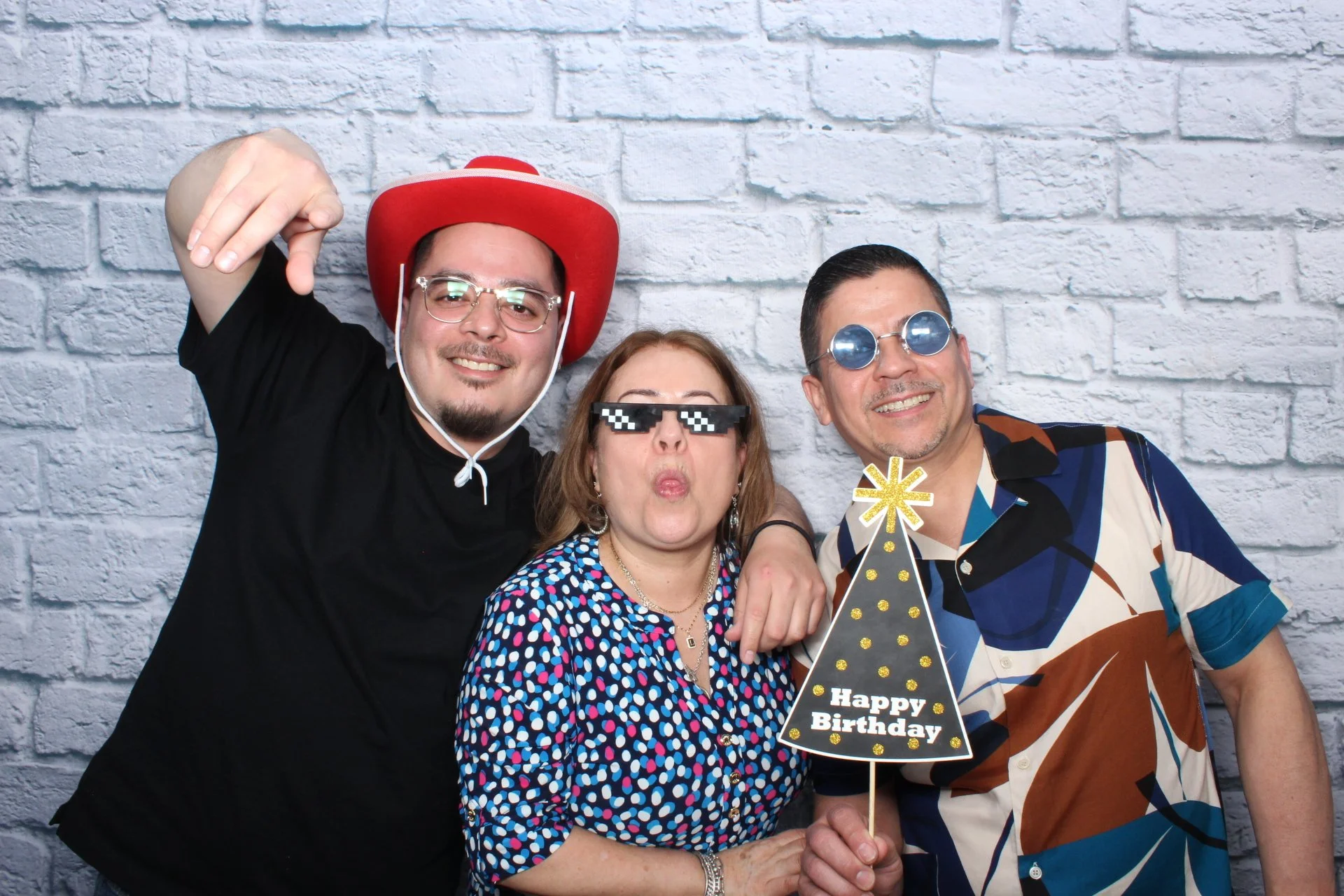Three friends celebrating a birthday, wearing festive accessories, holding a Birthday hat decoration, standing in front of a white brick wall, smiling and posing for the camera.