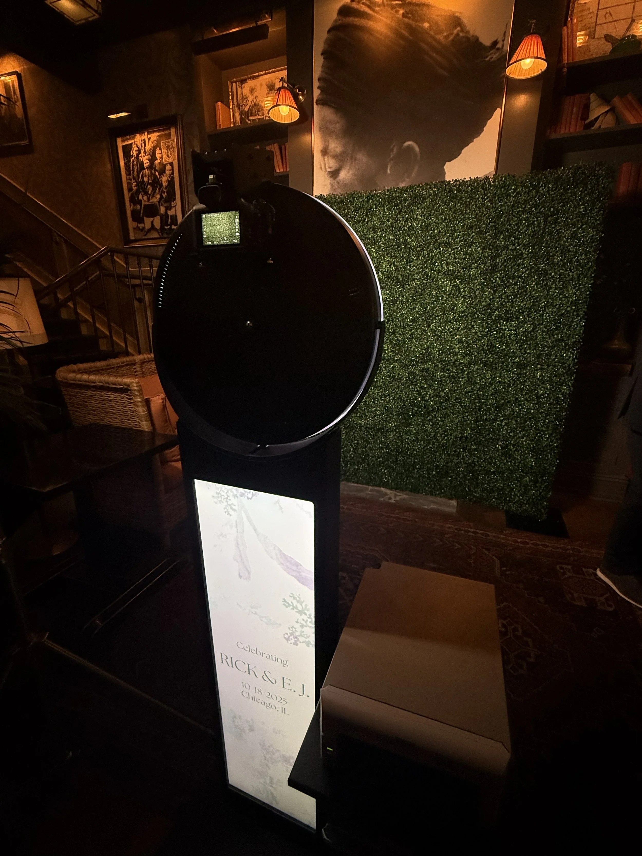 Photobooth setup with a large decorative photo of a person with a top knot hairstyle, a smaller framed picture of a group of people, and a tall sign celebrating Rick & E.J. with event details, in a dimly lit indoor space with artwork, books, and warm