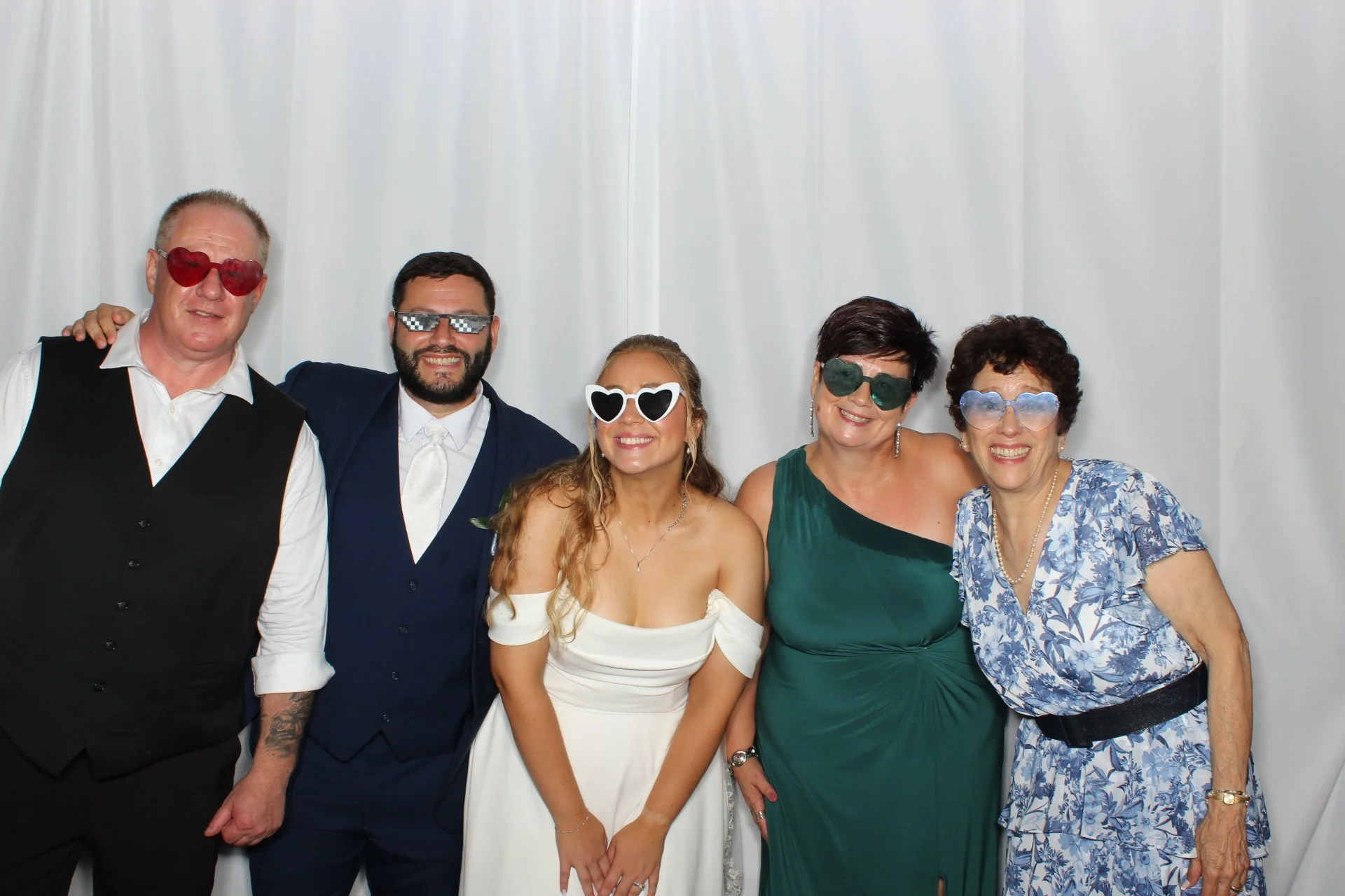 A group of five people dressed in formal attire, wearing fun sunglasses, posing together in front of a light-colored curtain at a celebration or event.