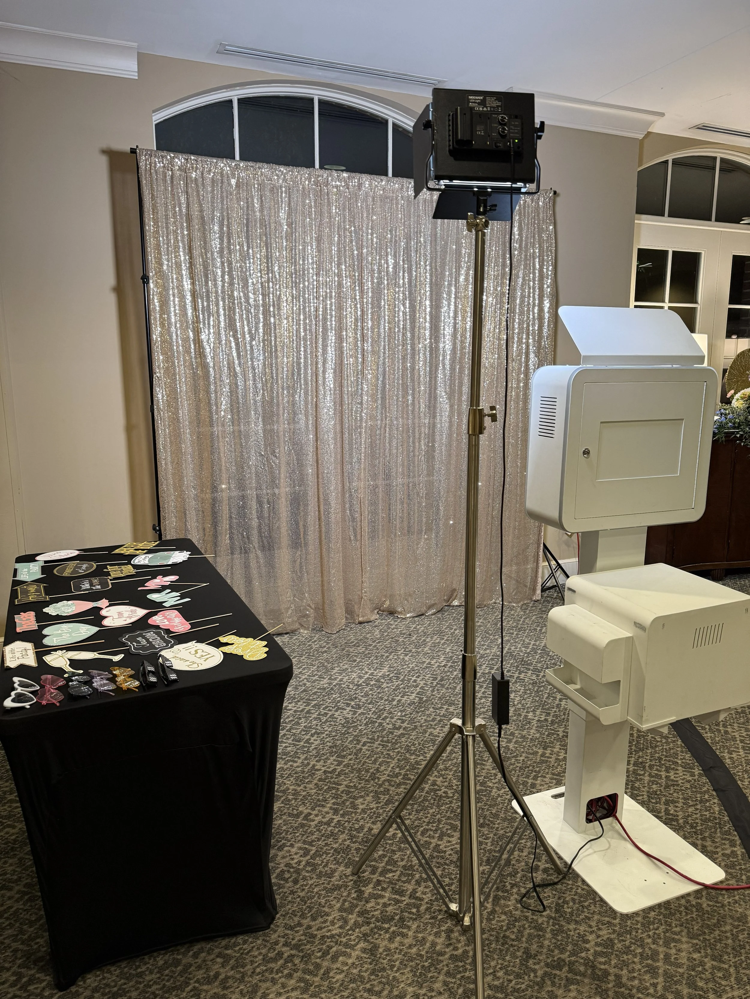Photo of a photo booth setup with a glittery backdrop, lighting equipment, a table with photo props, and photo booth printers on a carpeted floor in a room with large windows.