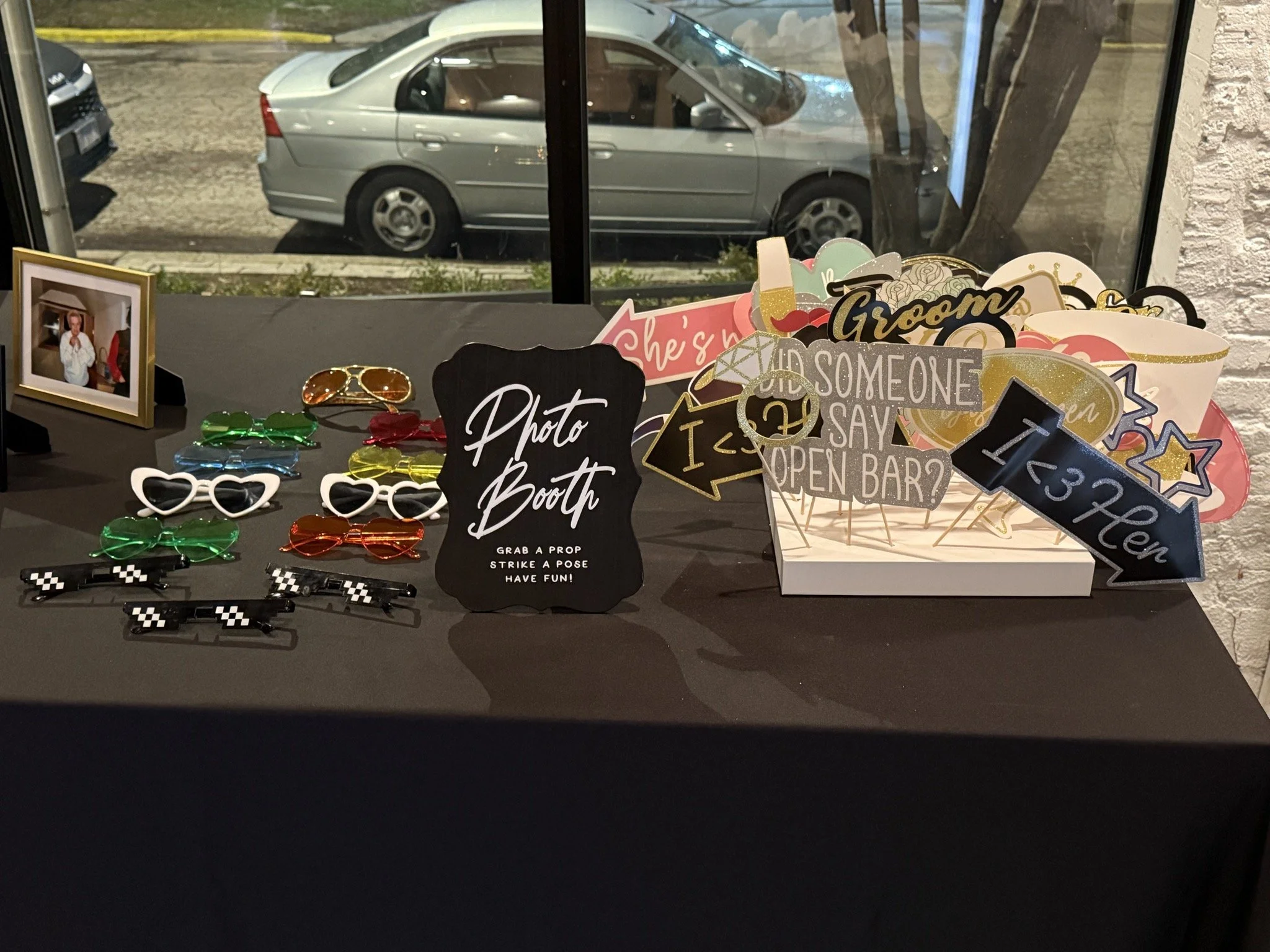 Photo booth table with various photo props, sunglasses, and picture frames, with a large window showing parked cars outside.
