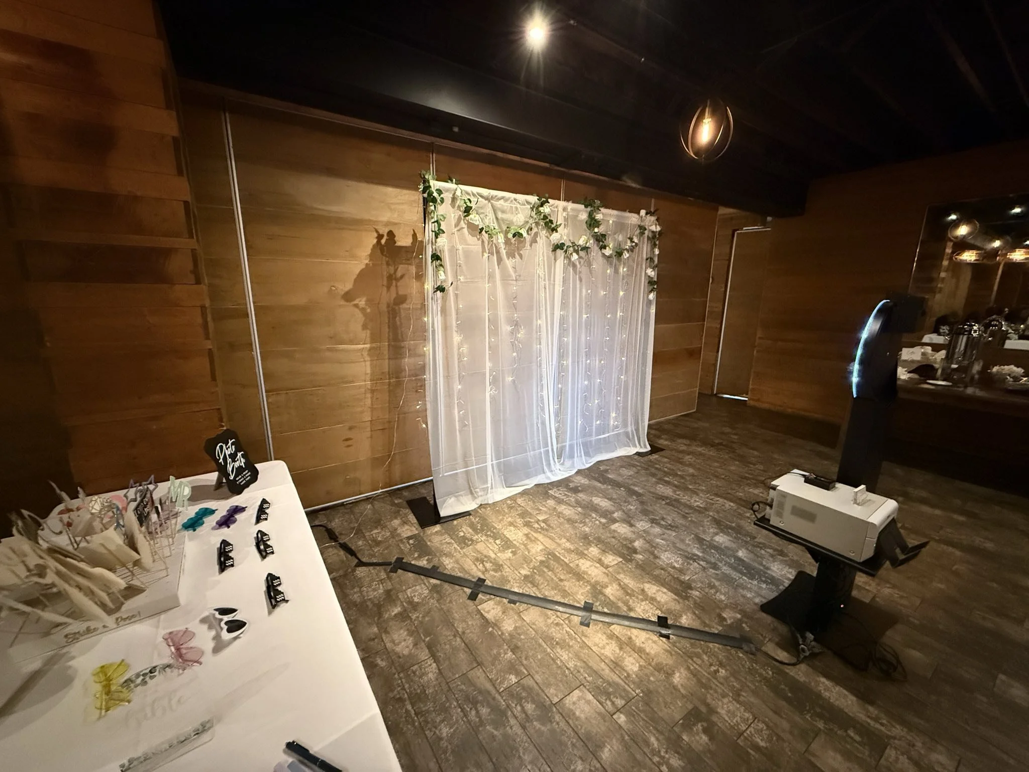 Party photo setup with a white sheer curtain decorated with string lights and greenery, and a table with various photo props and accessories in a room with wooden walls and ceiling, and a projector.