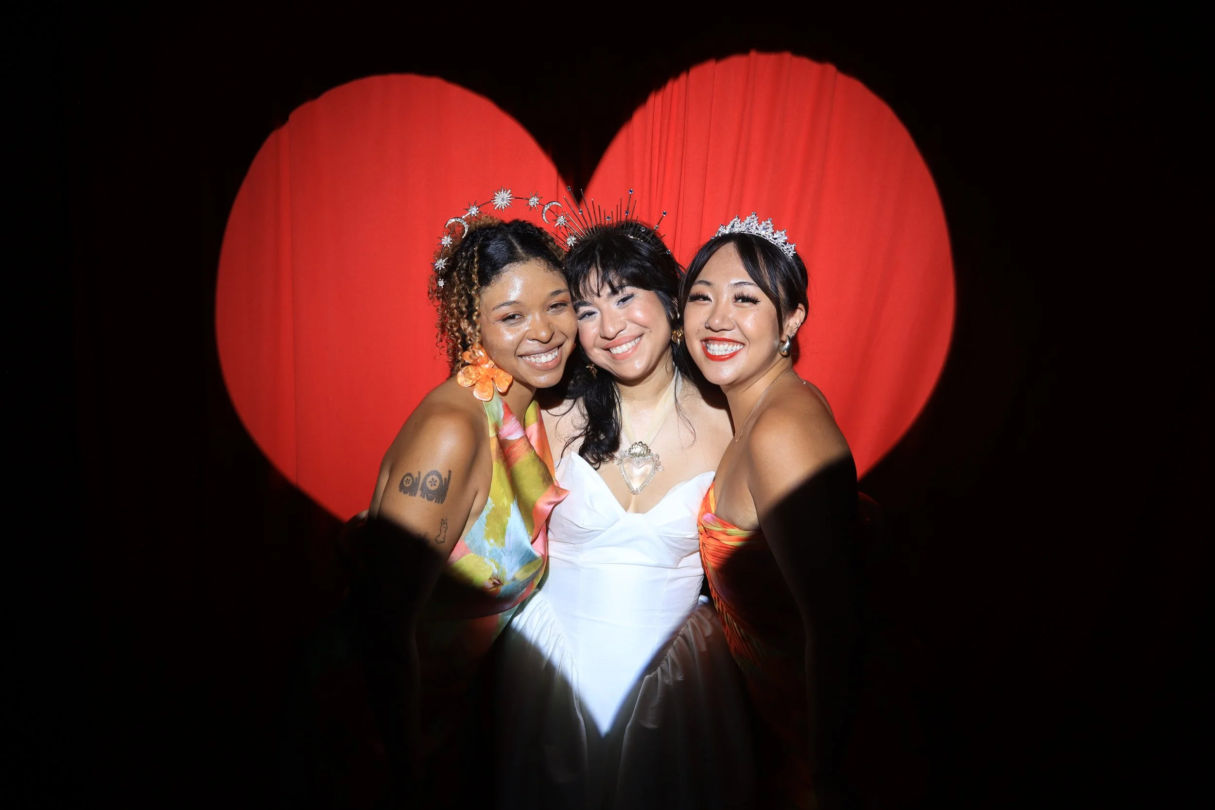 Three smiling women wearing tiaras taking a group photo in front of a large red heart backdrop.