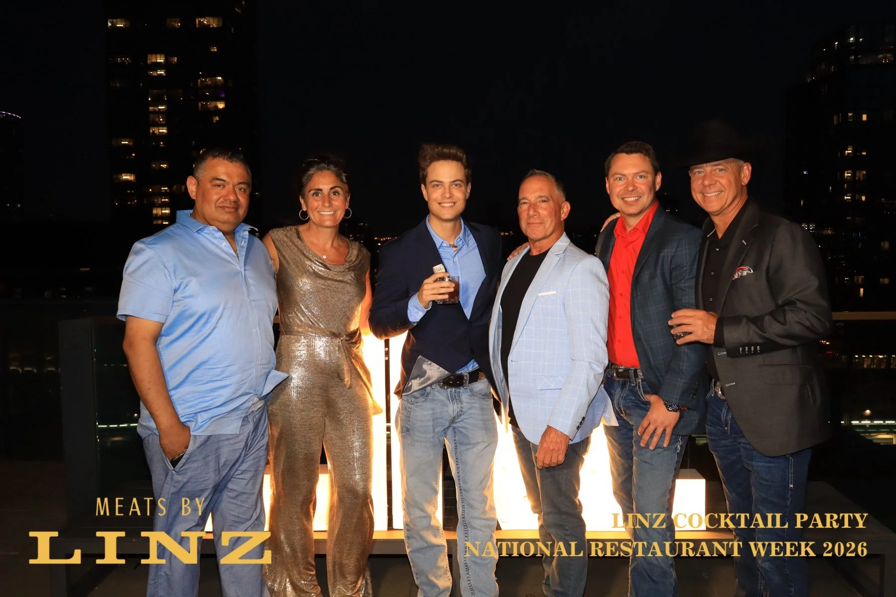Group of six people at a cocktail party on a rooftop at night, with city lights in the background. They are dressed in semi-formal to casual attire, smiling, some holding drinks. The event is part of the Linz Cocktail Party during National Restaurant