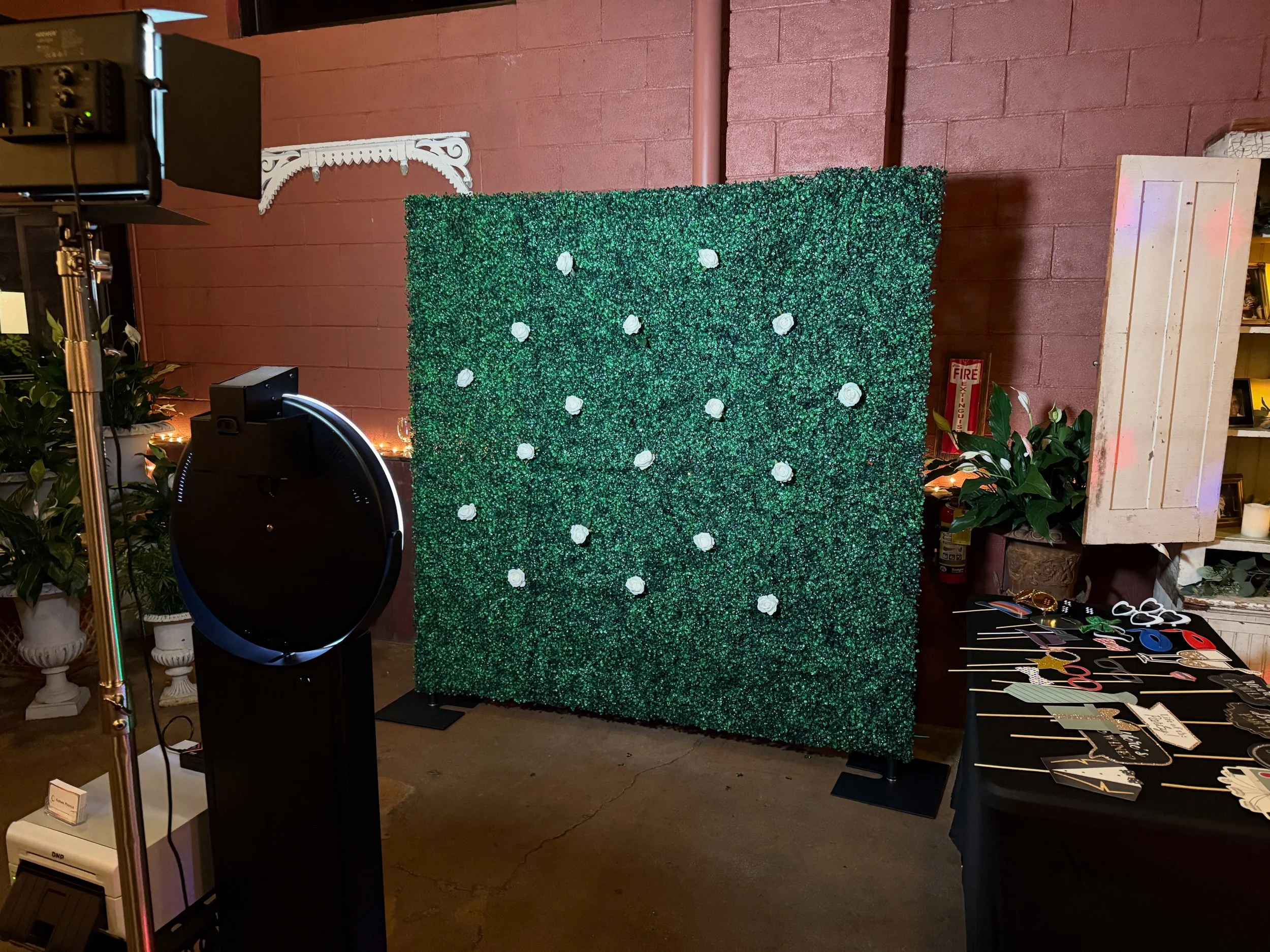 A decorated photo backdrop made of green foliage with white roses, arranged for an event. To the right, there are photo booth props like cutouts and signs on a table. To the left, photo booth equipment is visible.