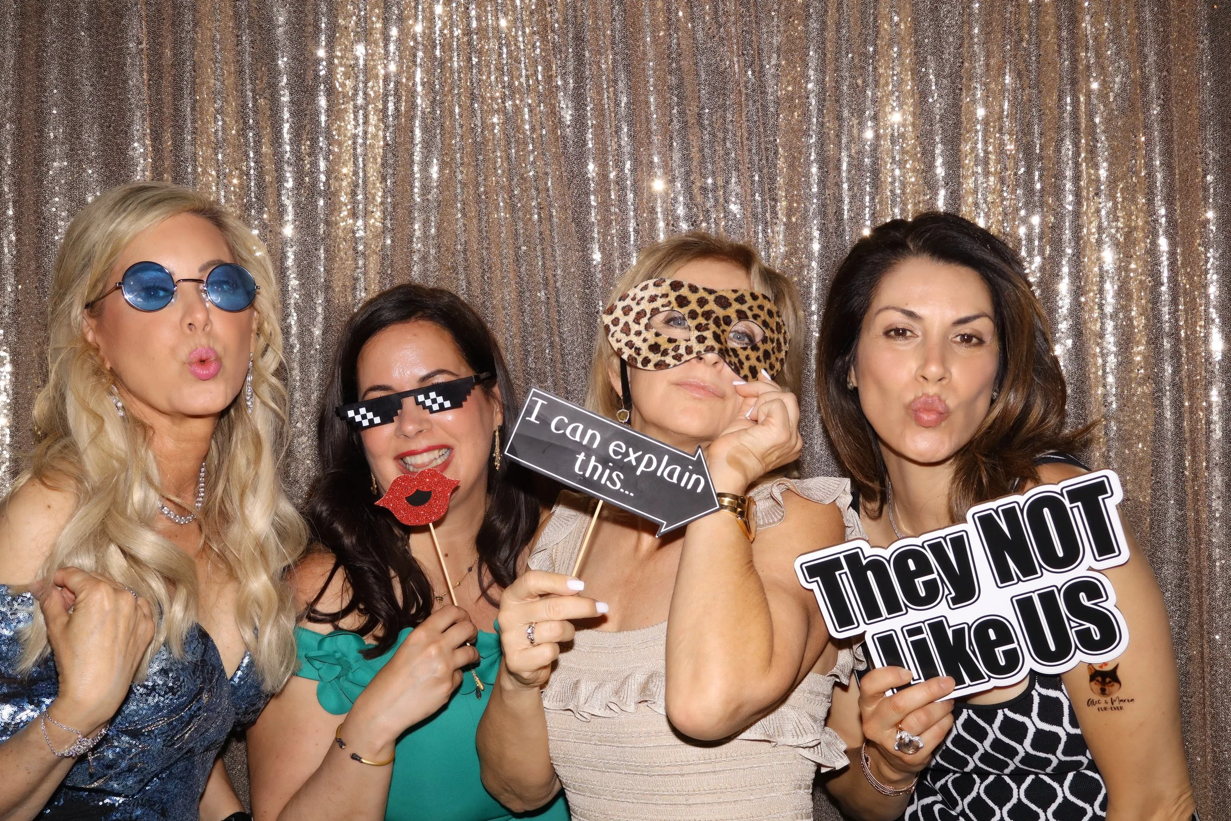 Group of women at a photo booth wearing fun accessories and holding signs, standing in front of a sparkly backdrop.