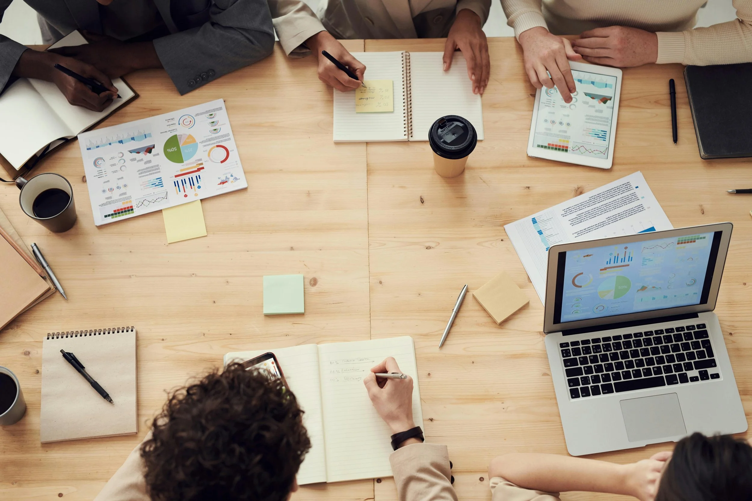 A top-down view of a group meeting at a wooden table with documents, notebooks, tablets, laptops, pens, paper cups, and sticky notes.