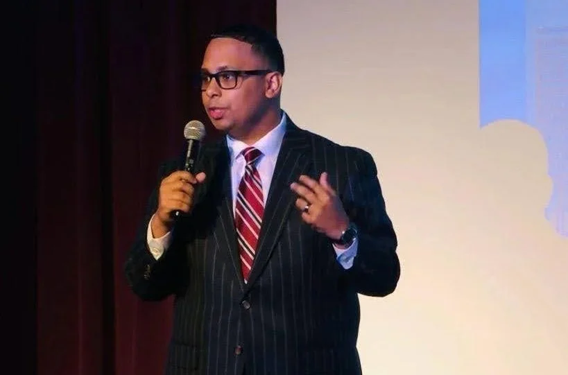 A man in a dark pinstripe suit with a red and white striped tie, black glasses, holding a microphone, speaking on stage with a presentation screen in the background.
