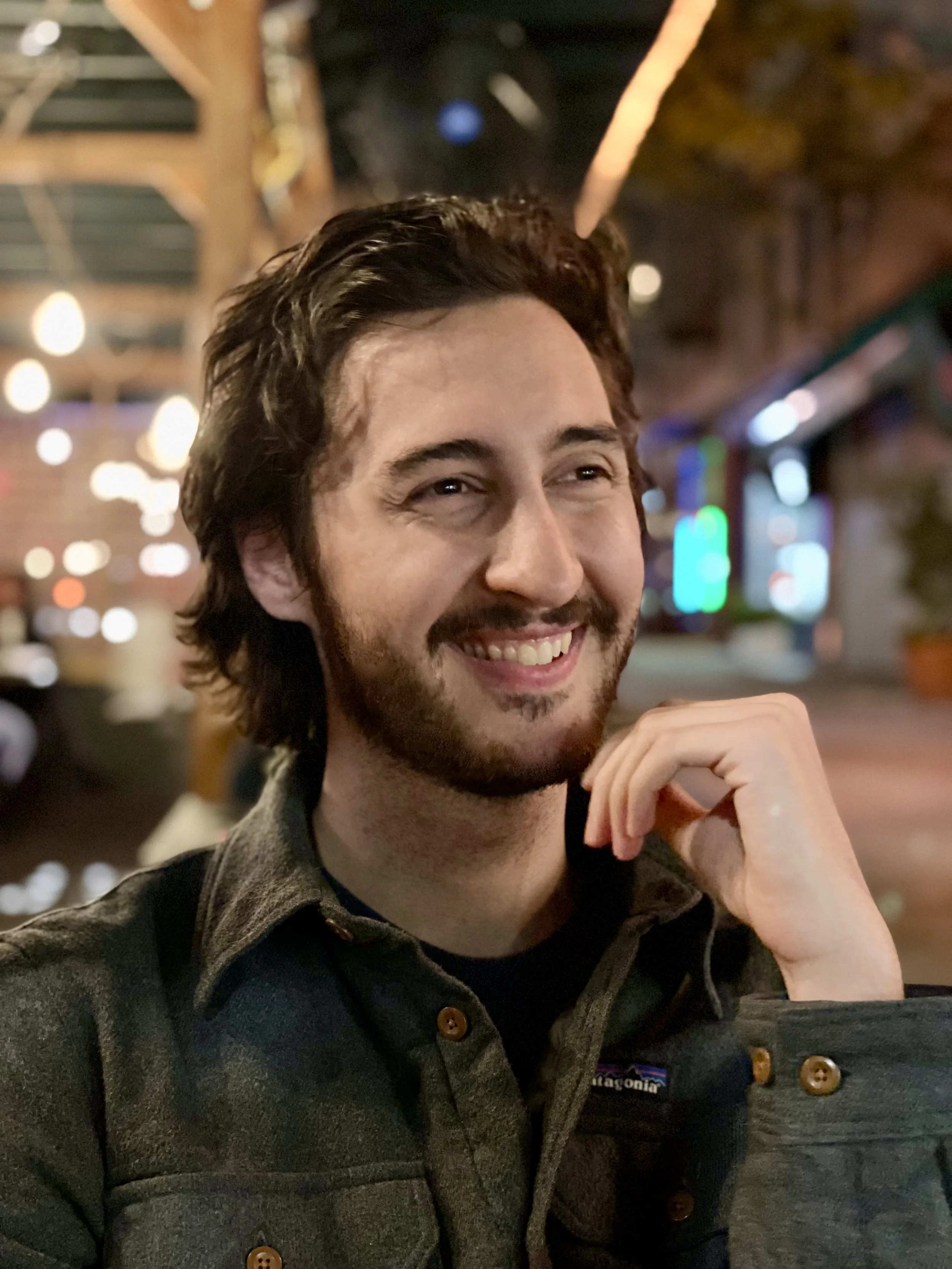 A smiling man with dark hair and a beard, wearing a gray flannel shirt, sitting in a cozy, warmly lit indoor or outdoor cafe with blurred colorful lights in the background.