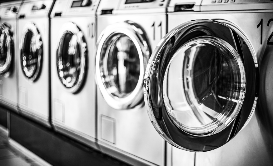 Black and white photo of a row of front-loading washing machines in a laundromat, with the closest one showing its empty drum.