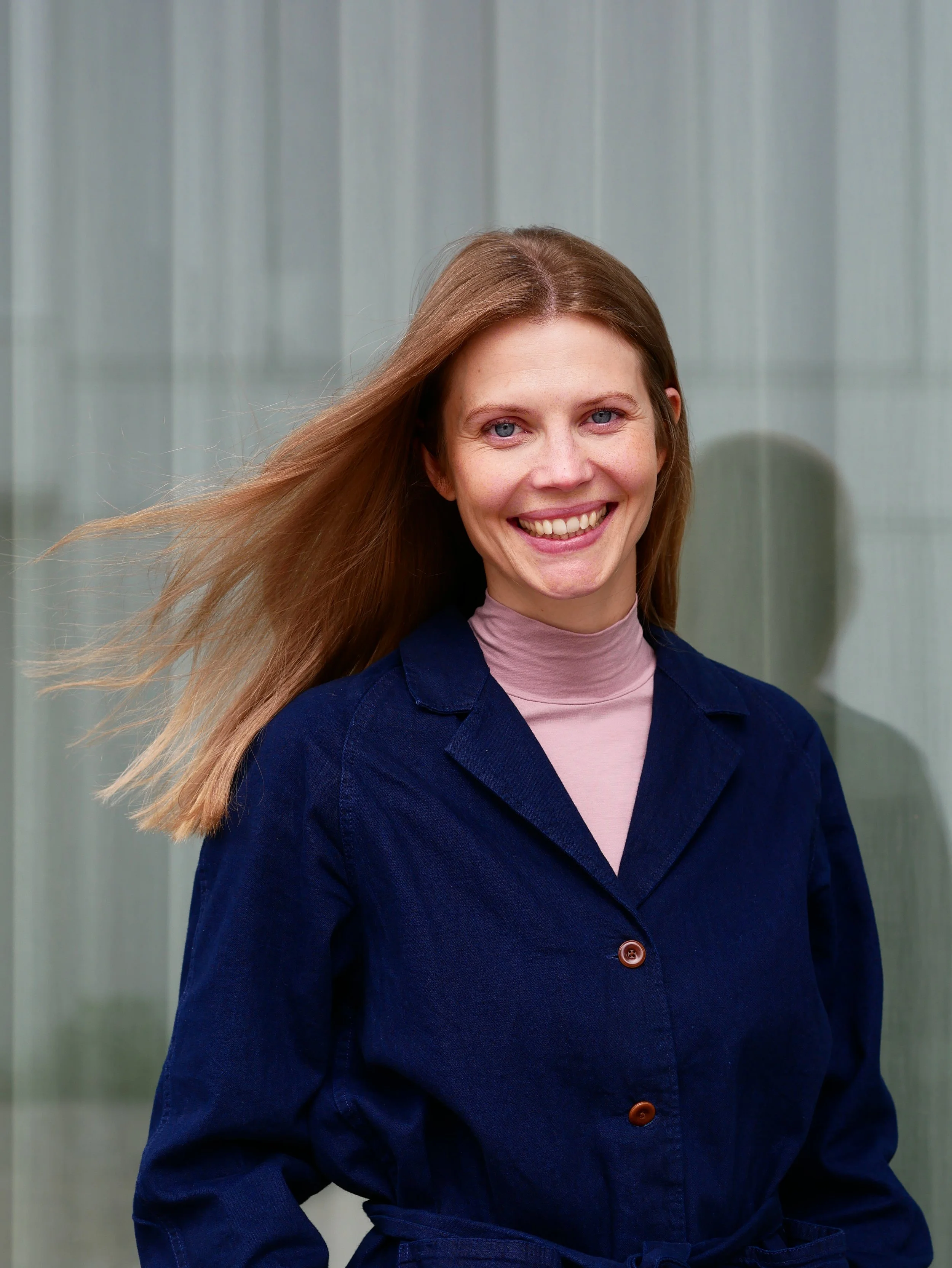 Junge Frau mit langem, braunem Haar, blauen Augen, im blauen Mantel und rosa Pullover, lächelnd vor einem transparenten, silbernen Hintergrund.