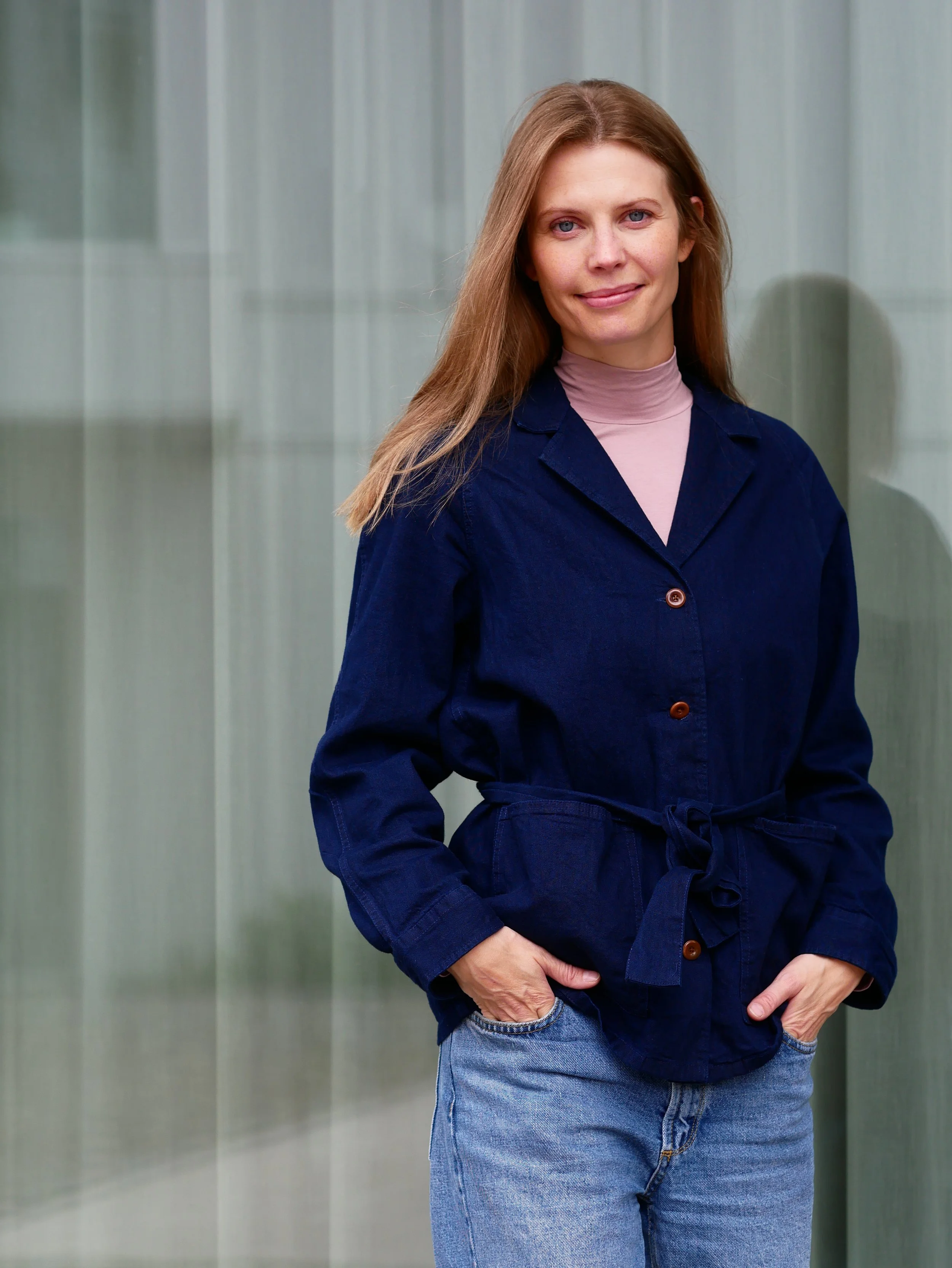 Eine Frau mit warmem Lächeln trägt eine dunkelblaue Jacke und eine rosa Bluse, ihre Hände sind in ihre Hosentaschen gesteckt, vor einem grauen Hintergrund.