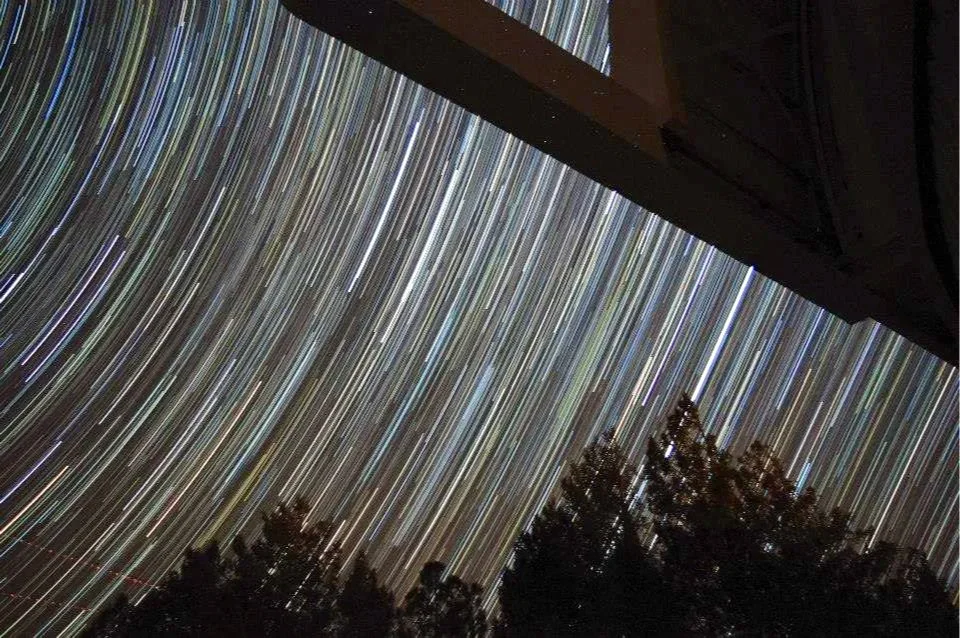 Star trails visible through the night sky with trees and building silhouetted in the foreground.