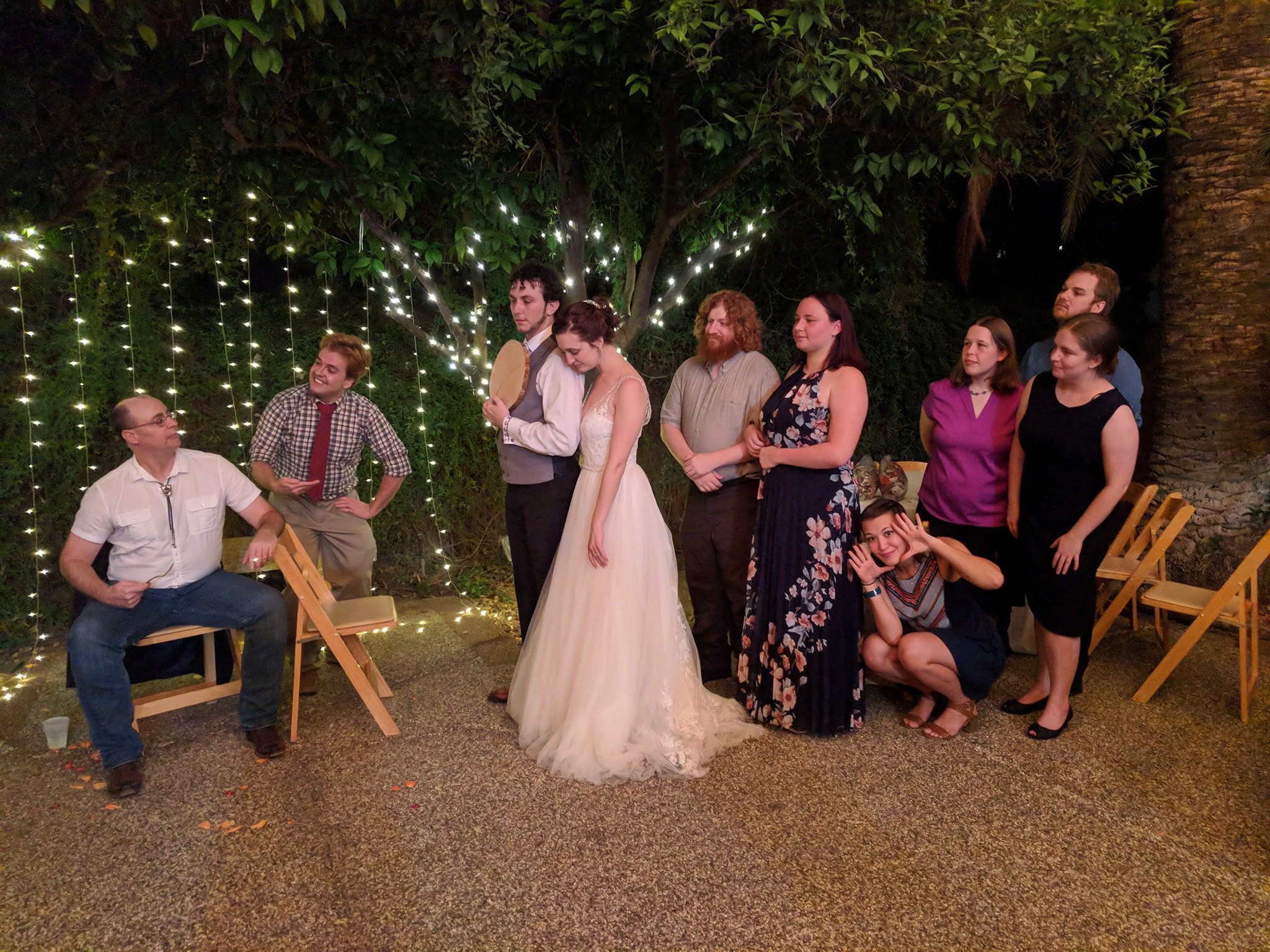 A group of people standing and sitting outdoors at night, under a tree decorated with string lights, at a wedding celebration.