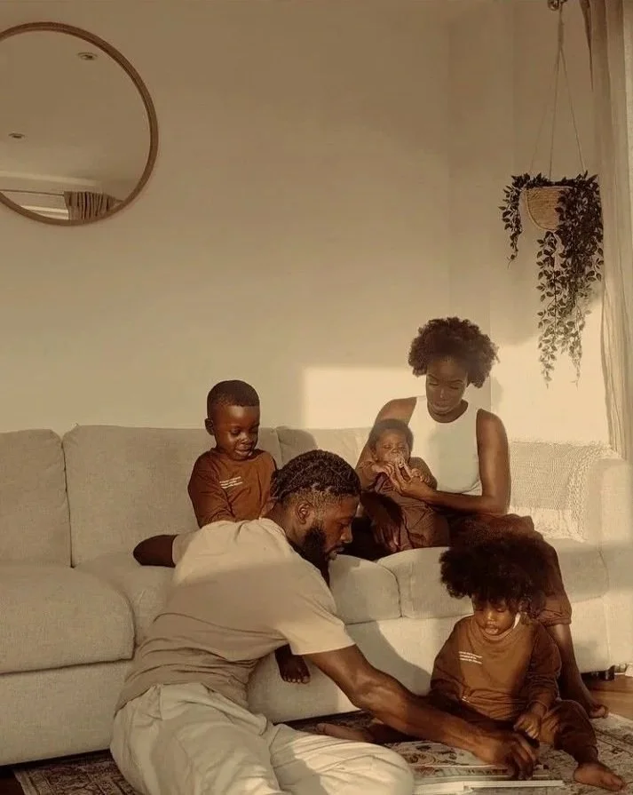 A black family with young children sitting together on a couch, playing and interacting in a warm space with sunlight.