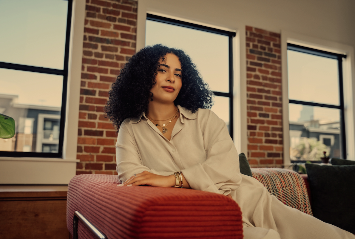 a Therapist, smiling in a warm office setting, with a orange couch and brick wall.