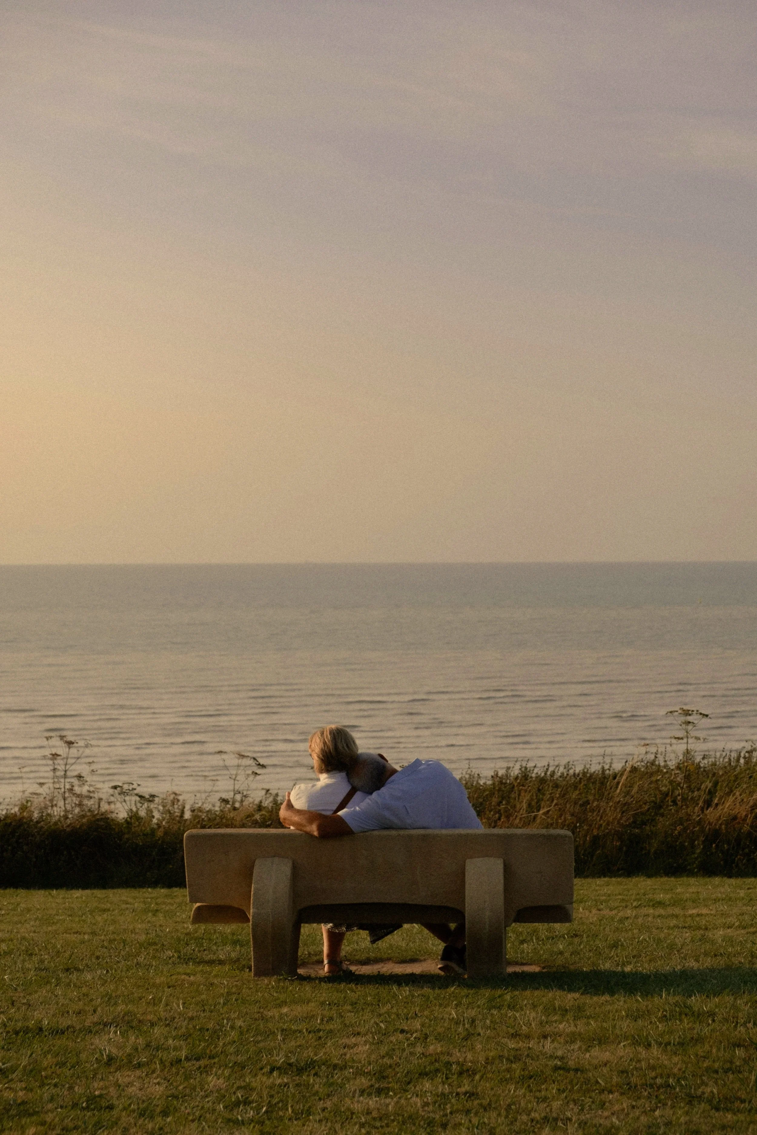 A couple embracing on a bench, facing the ocean together, in a peaceful setting.