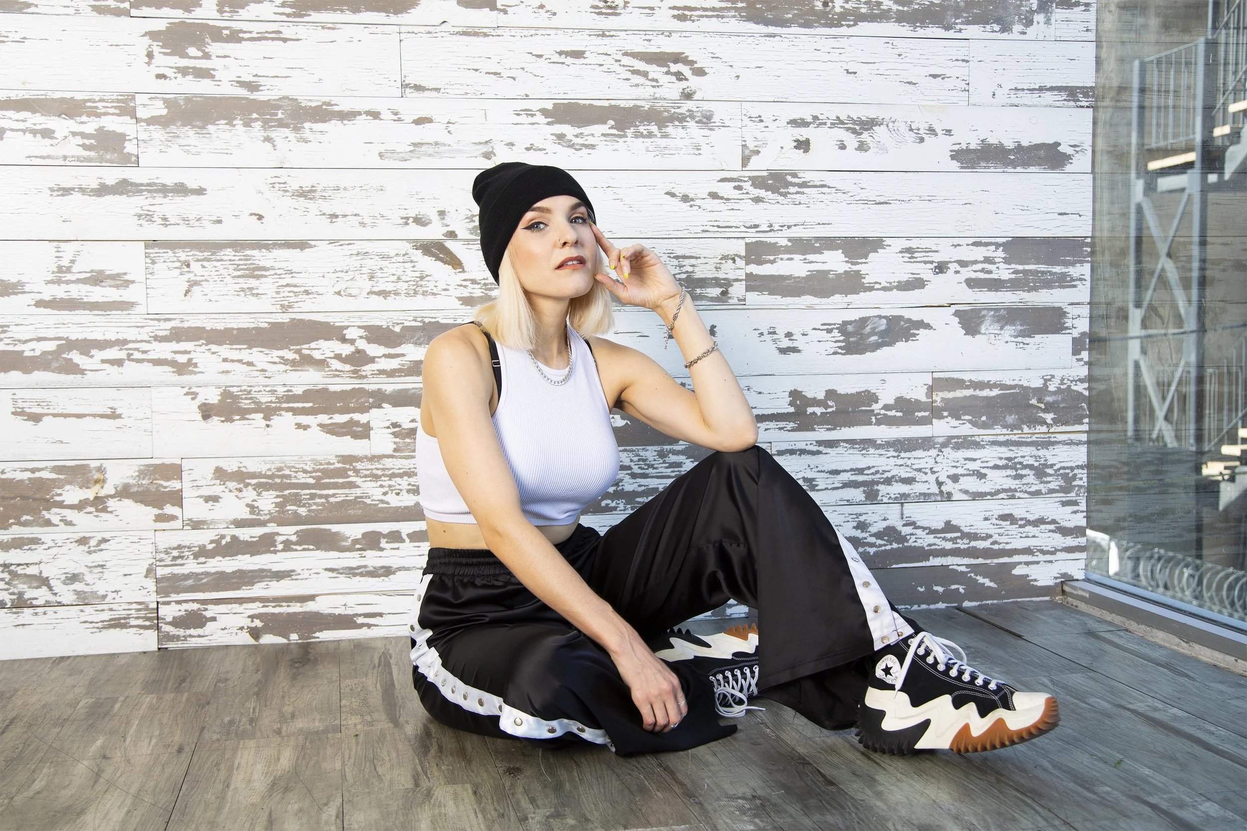A blonde model sits in an unfinished studio space wearing a black and white streetwear look, including striped black track pants, a white cropped tank top and a black beanie hat. 
