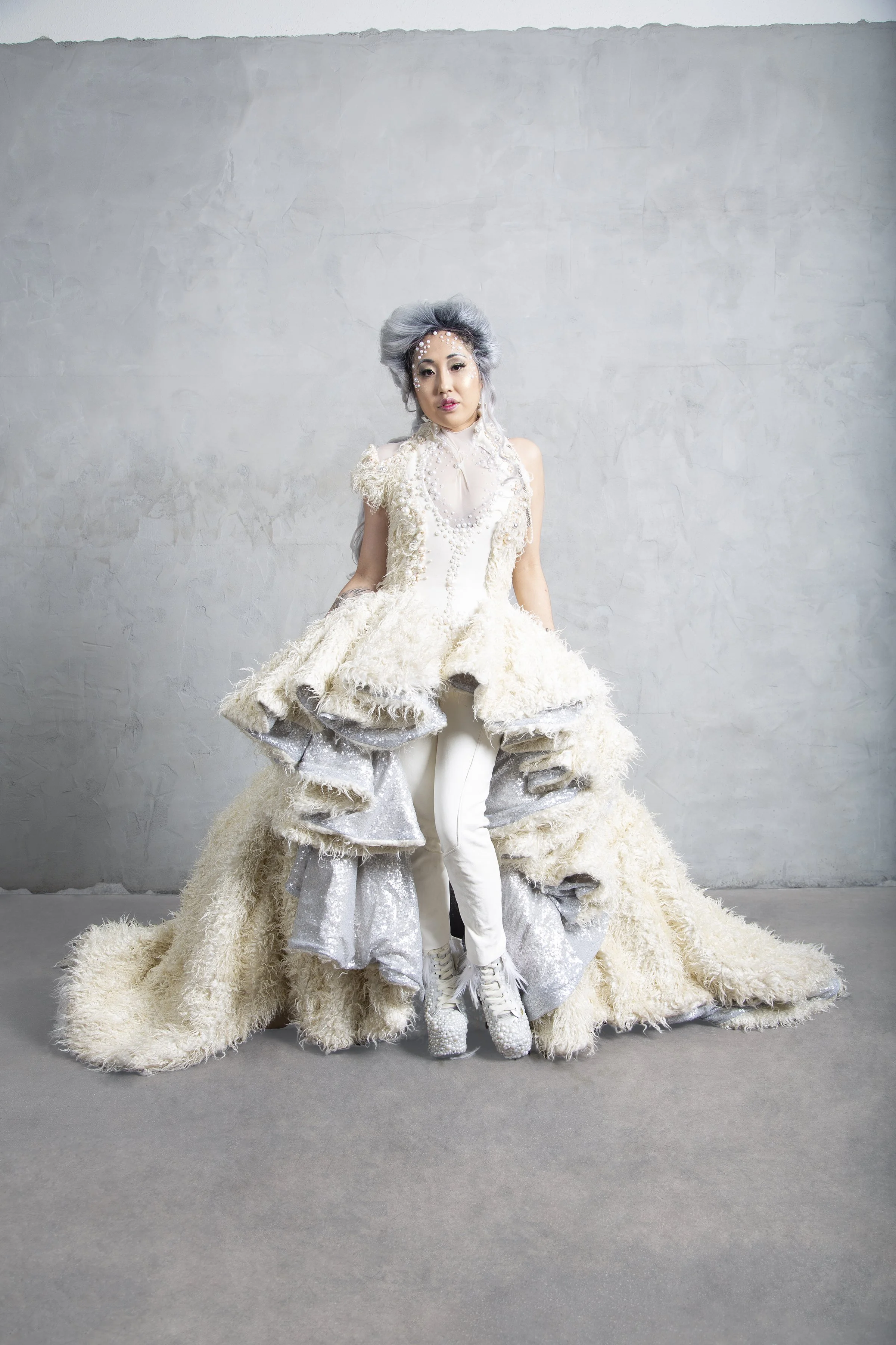 Set in a studio, an Asian American model wearing a custom couture dress featuring layered panels and a long train. She is wearing a grey Elizabethan style wig.