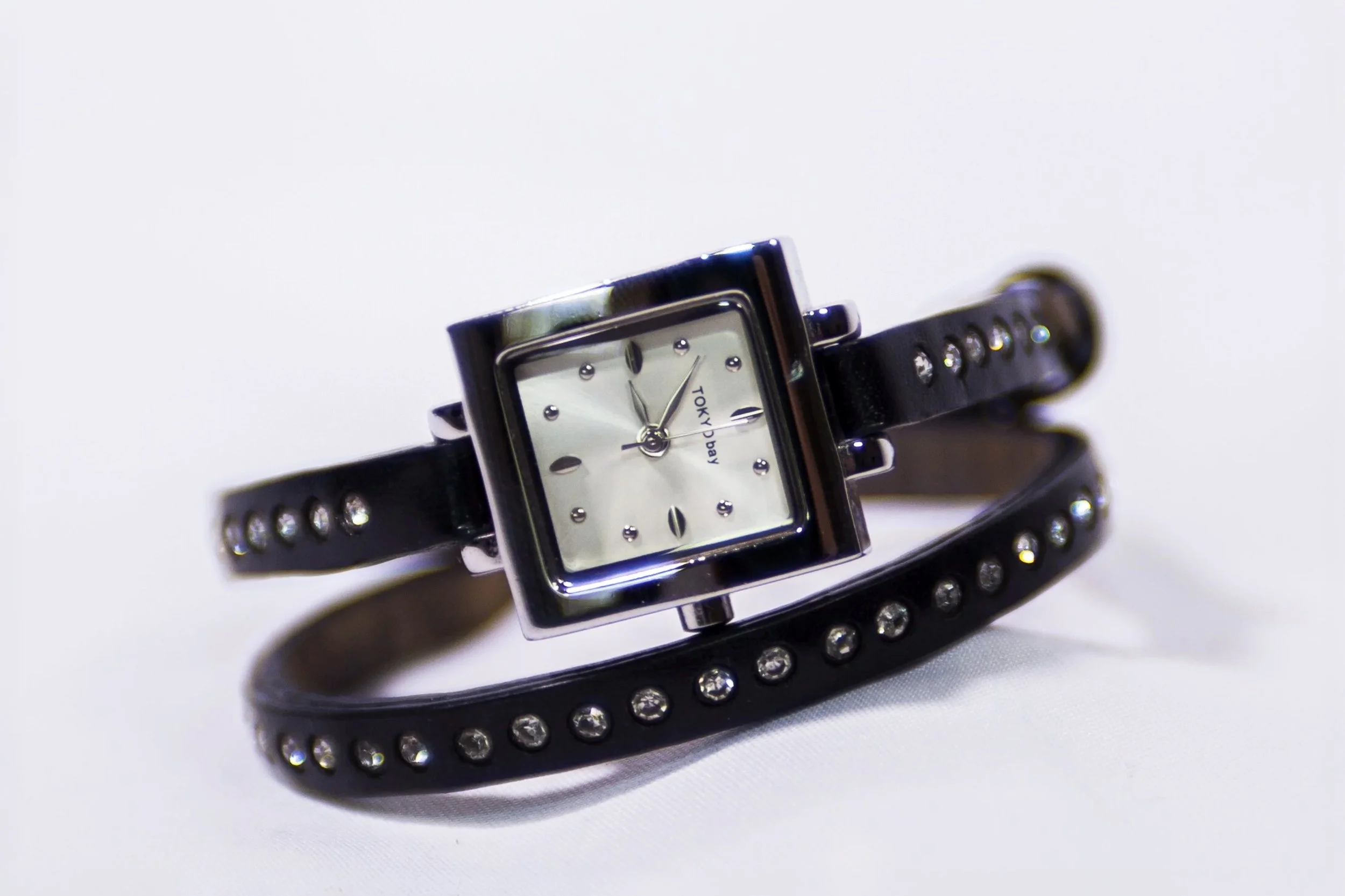 Product shot of a Tokyobay wrist watch on white background. Watch face features pearl and silver numbering. Wrist strap is a double wrap black leather band with rhinestone embellishment. 