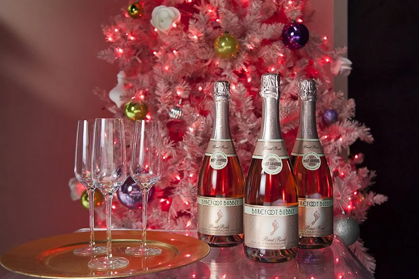 Still life editorial product shot of Barefoot Wines Sparkling Rosé wine. Three bottles on a glass table in front of a pink Christmas tree with a tray holding three Champagne flutes. 