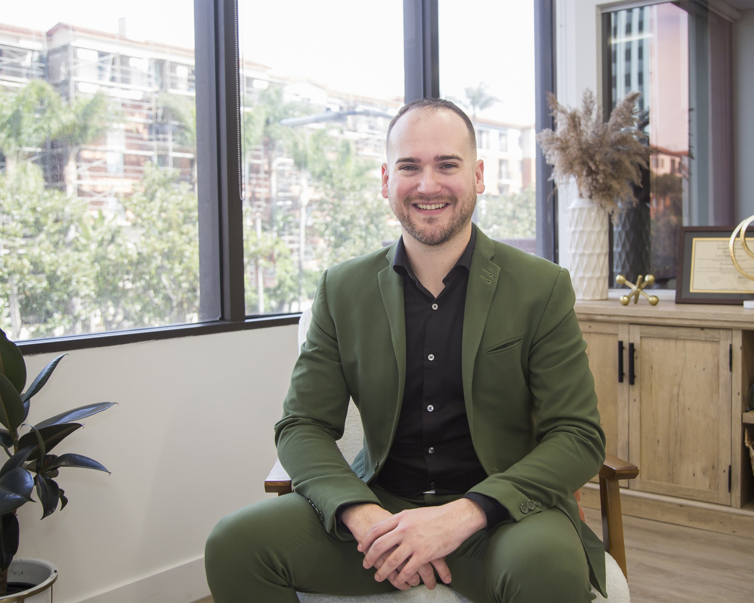 Executive portrait of a man wearing a green suit in an office, sitting in front of a large window. 