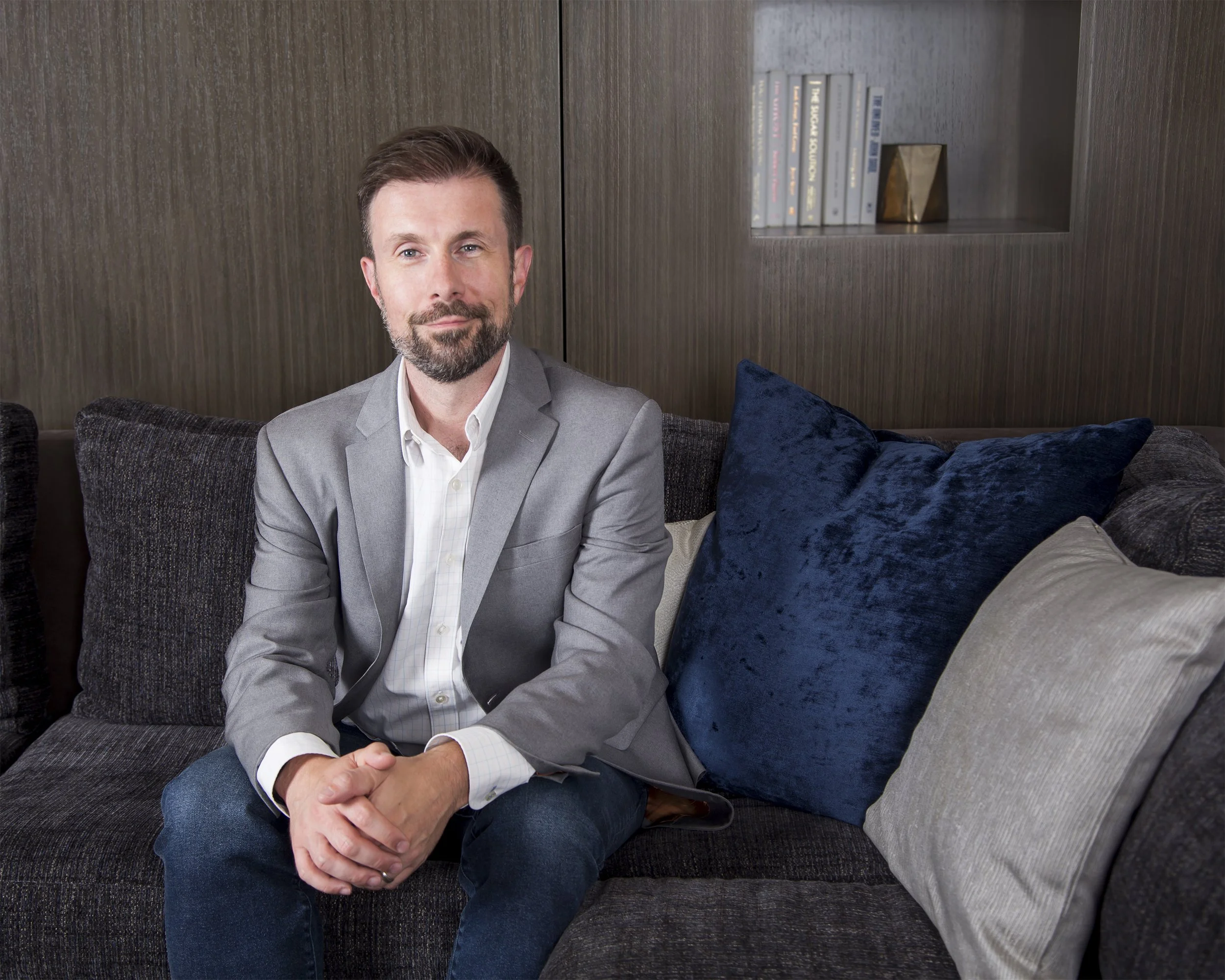 Executive portrait of a man with a beard sitting on a couch with books behind him. 