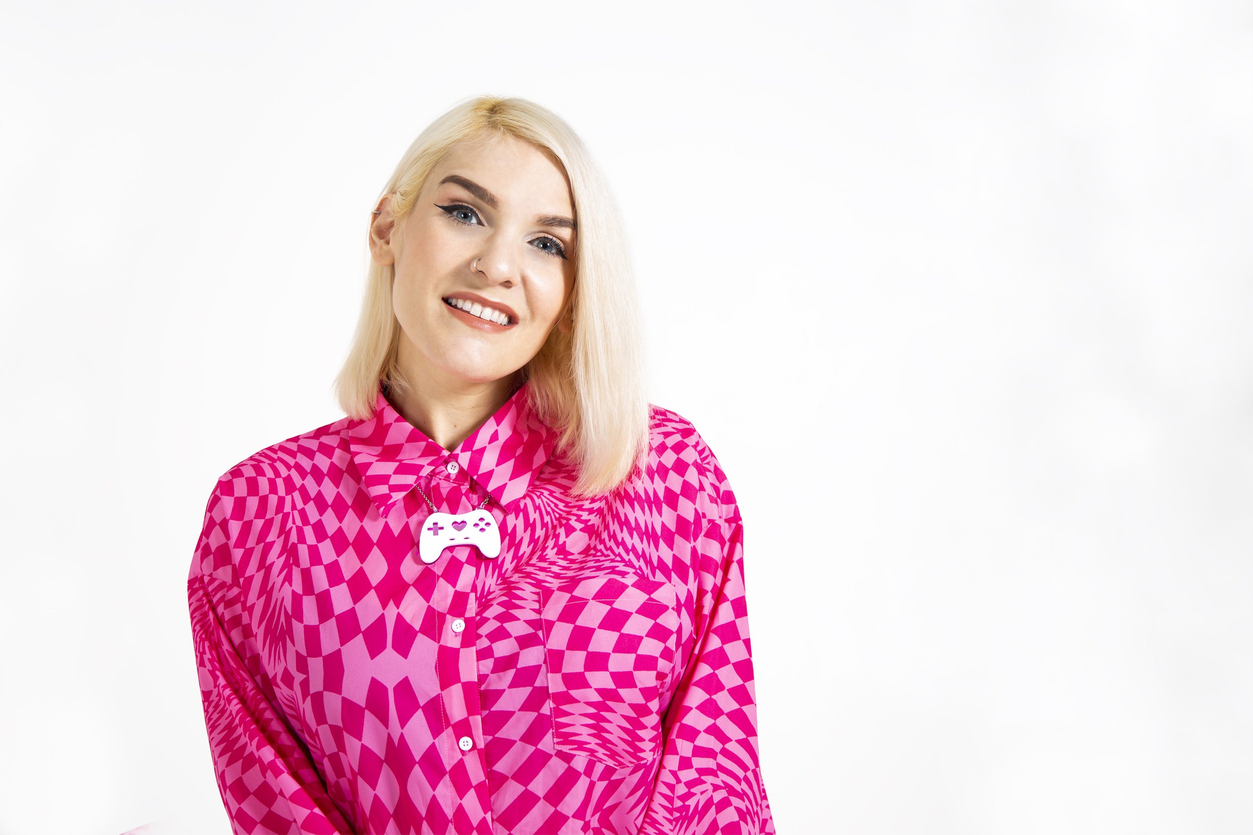 A blonde woman smiles wearing a pink top and gamer necklace on a white background.