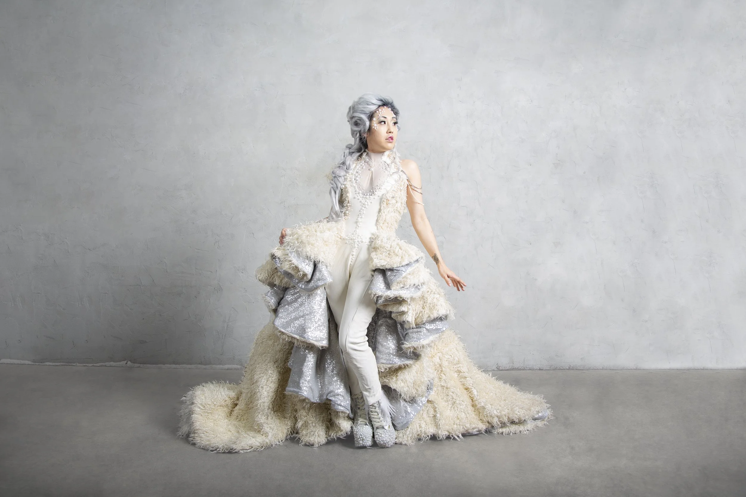 Set in a studio, an Asian American model wearing a custom couture dress featuring layered panels and a long train. The dress is inspired by the Luck Dragon, Falkor, from the movie The Neverending Story. She is wearing a grey Elizabethan style wig.