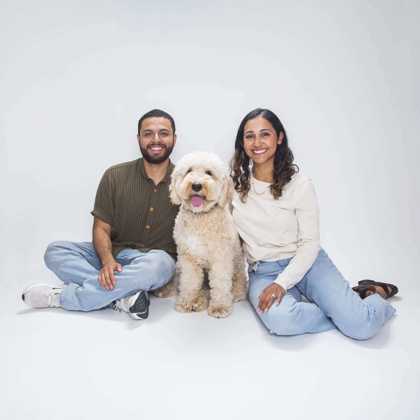 A Latino couple sits with their white doodle dog in the studio on a white background for their family portrait.