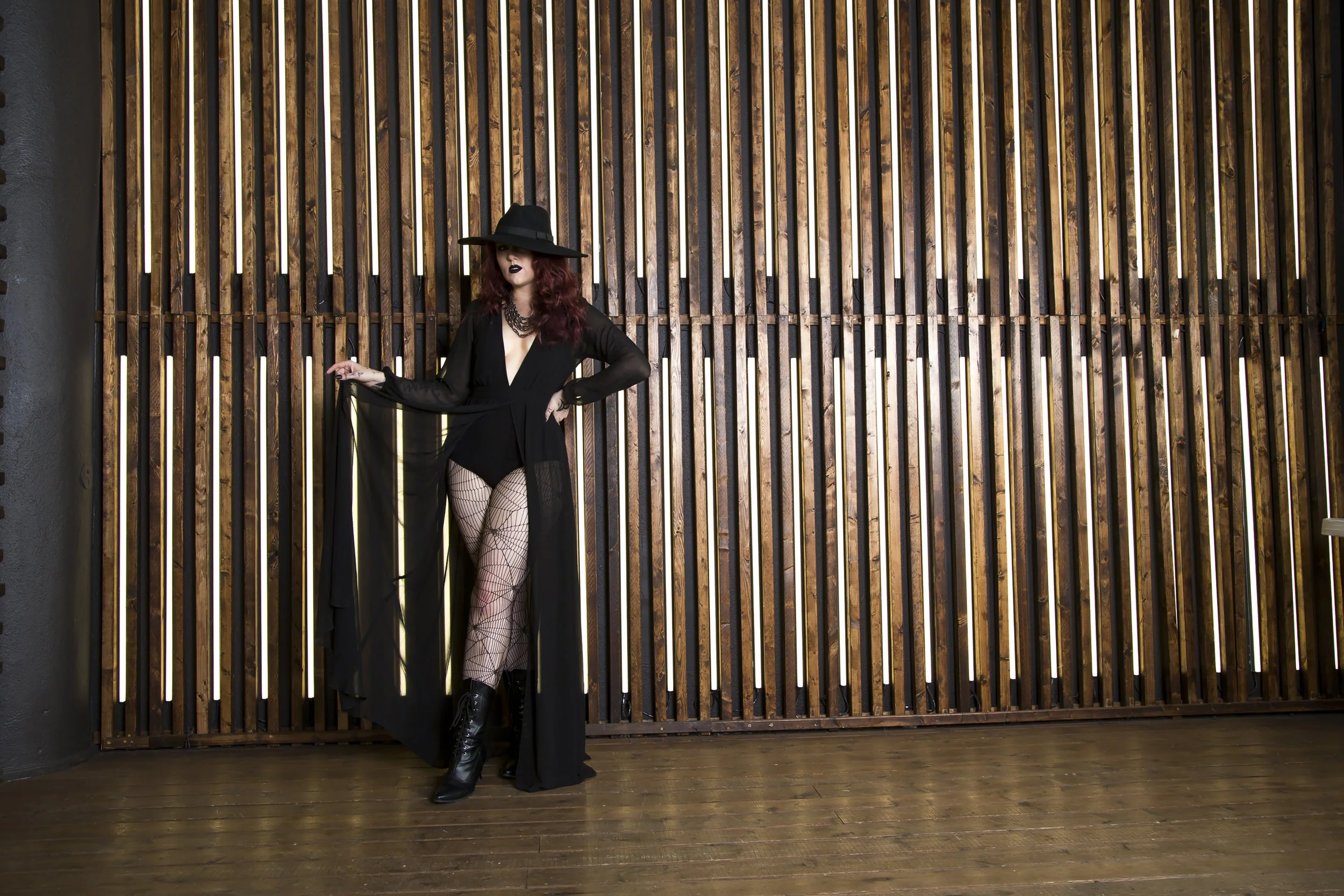 A tall red haired model wearing a wide brimmed hat and an all black high fashion witch look stands in front of a wooden wall with light coming through the slats.