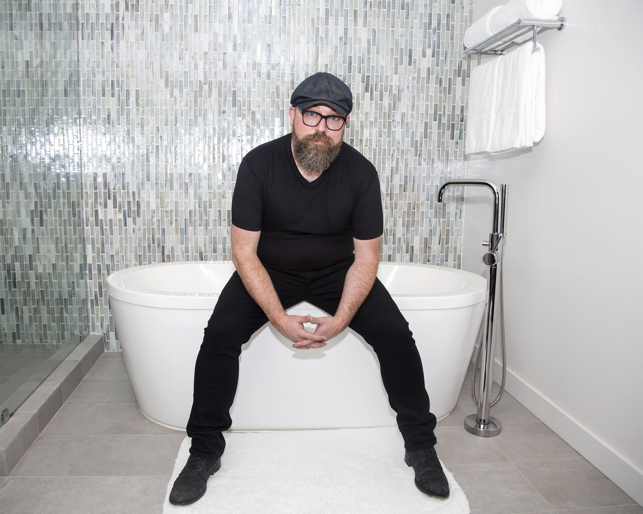 A man wearing a cap and all black sits in the bathroom of a hotel room on the edge of a large bathtub. 