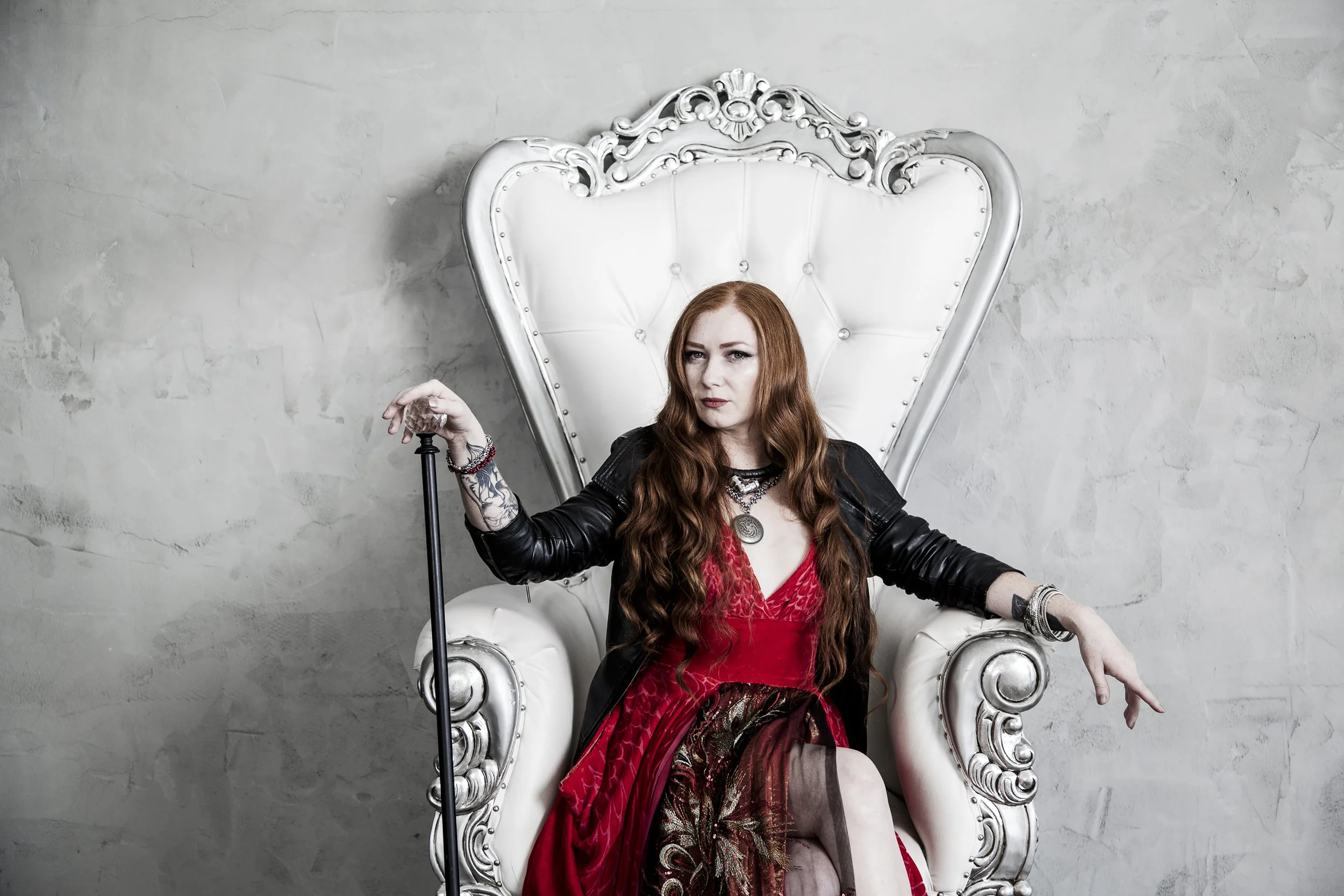 A red haired model sits in a white throne like chair in a studio. She is wearing a one of a kind red dress with peacock feather and a deeper red ombré hem.