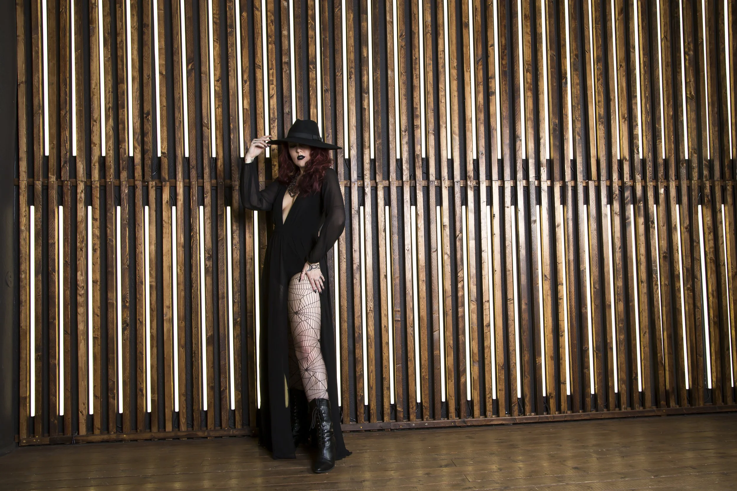 A tall red haired model wearing a wide brimmed hat and an all black high fashion witch look stands in front of a wooden wall with light coming through the slats.