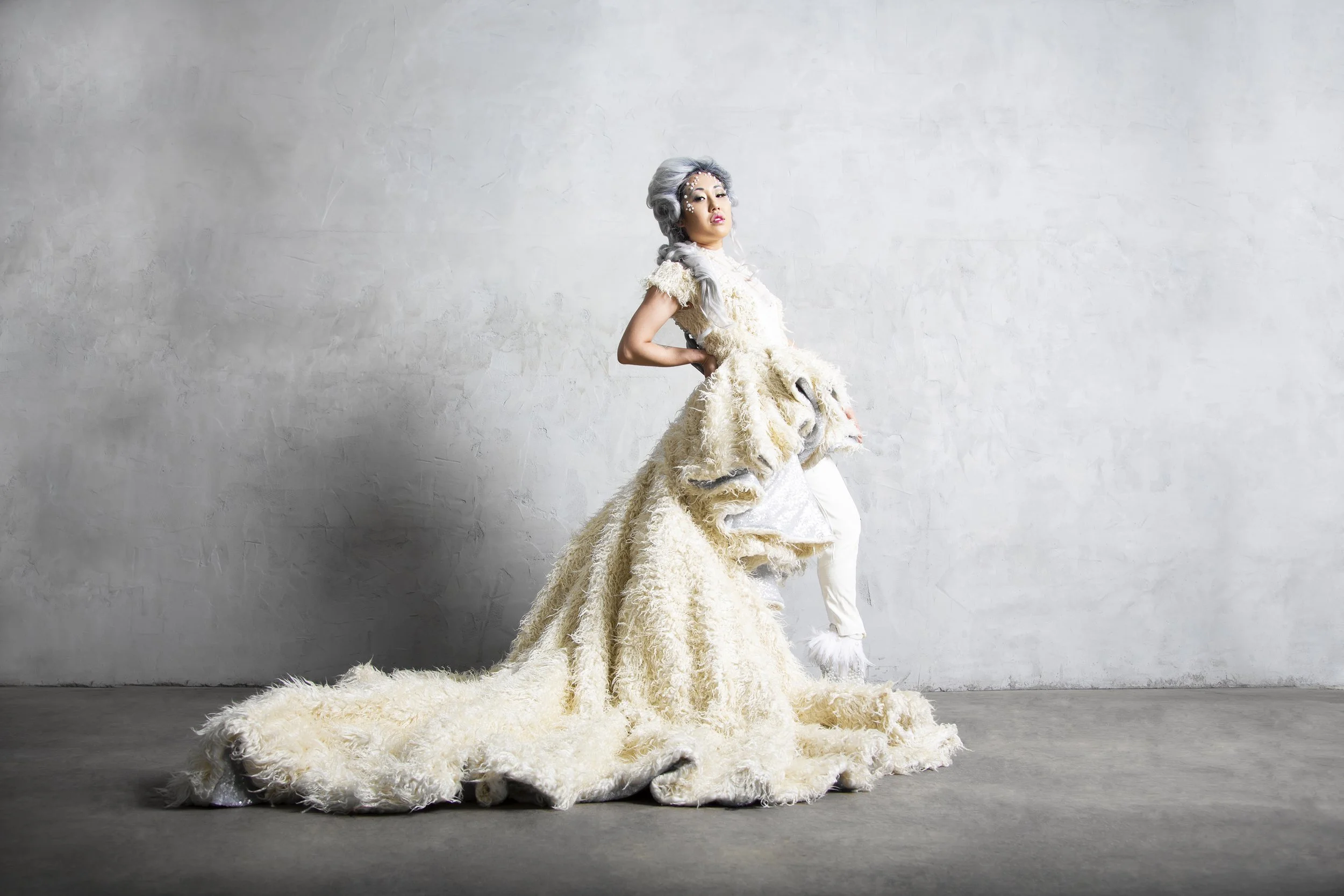 Set in a studio, an Asian American model wearing a custom couture dress featuring layered panels and a long train. She is wearing a grey Elizabethan style wig.