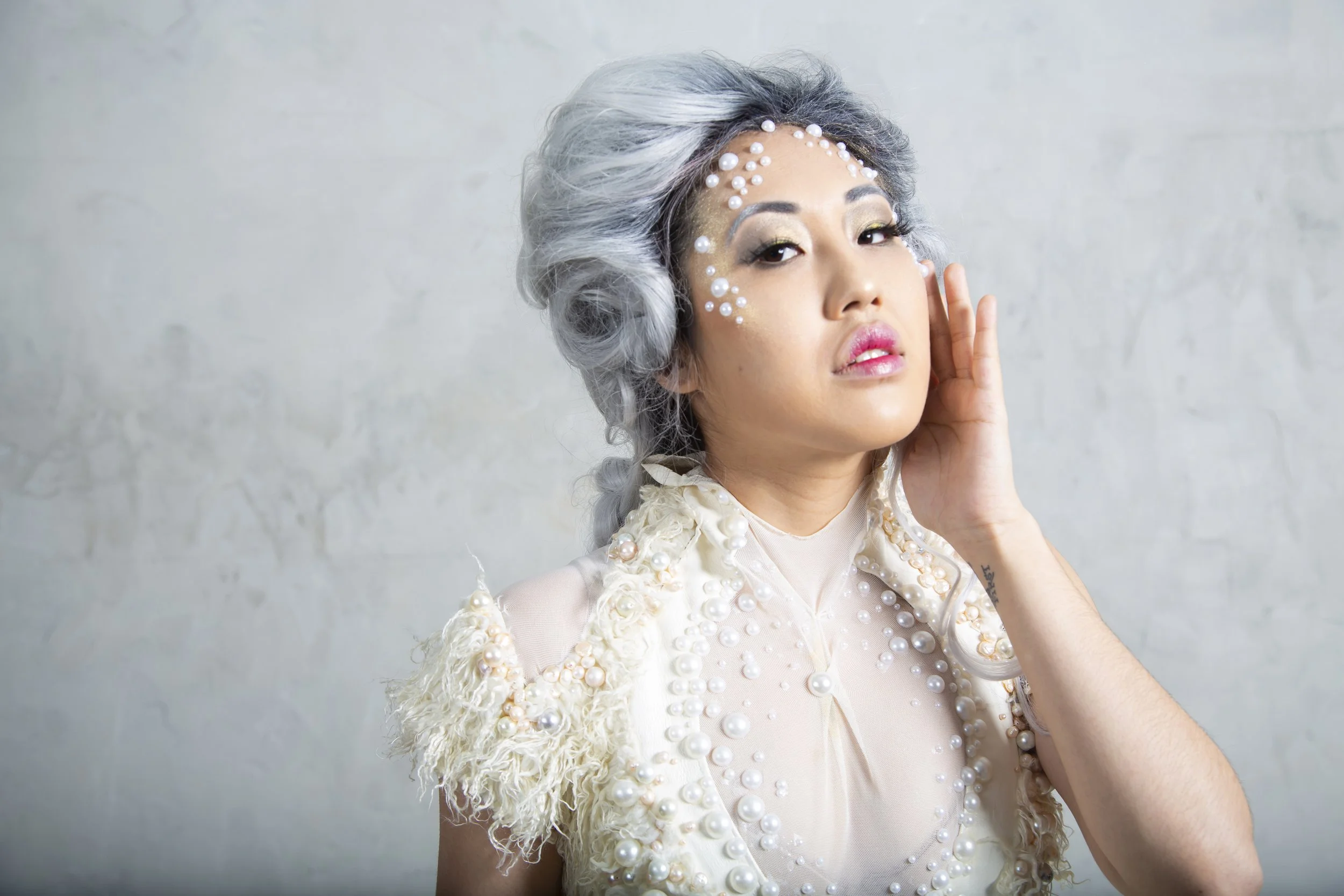 Set in a studio, close up image of an Asian American model wearing a custom couture dress featuring layered panels and a long train. She is wearing a grey Elizabethan style wig and has pearl details in her makeup.