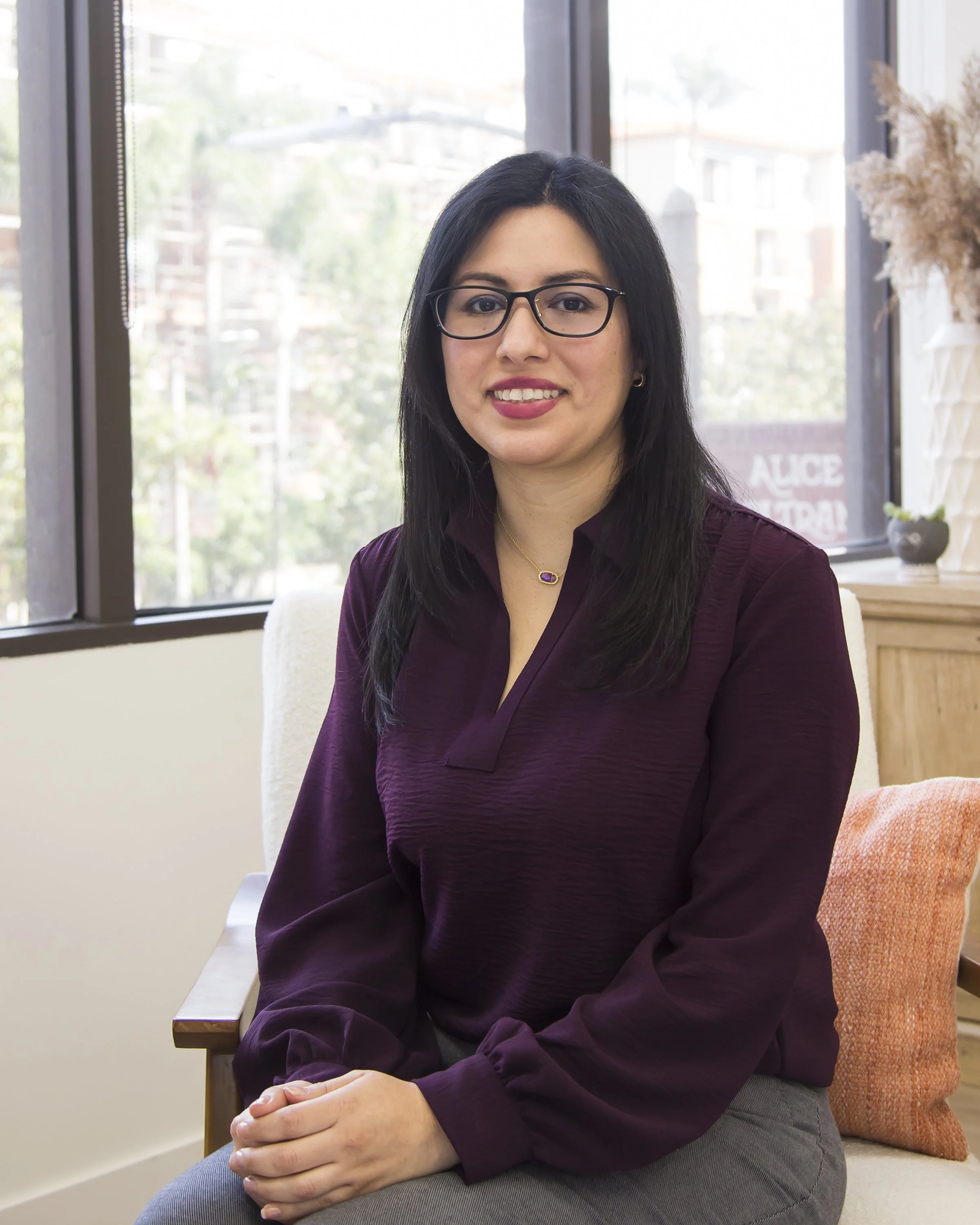 A Latina woman sitting in her office in front of a large window. She is wearing a purple top and glasses.