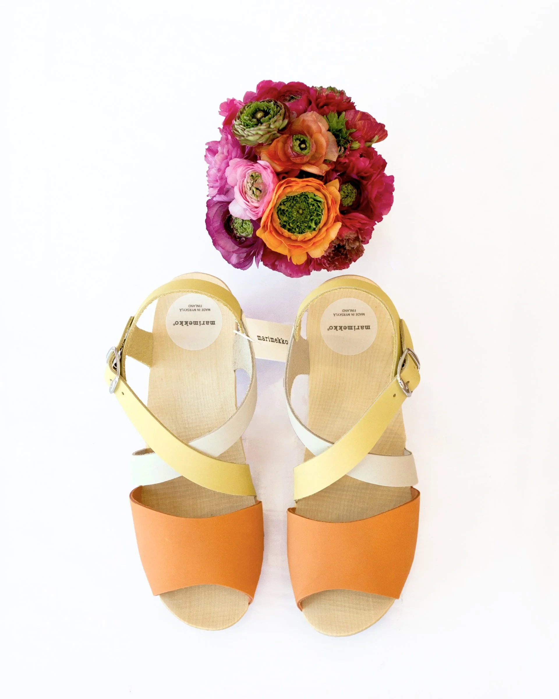 E-commerce product image of a pair of orange, white and yellow sandals with a wooden sole by Marimekko. There is a bouquet of flowers featured next to the shoes. The image is on a white background. 