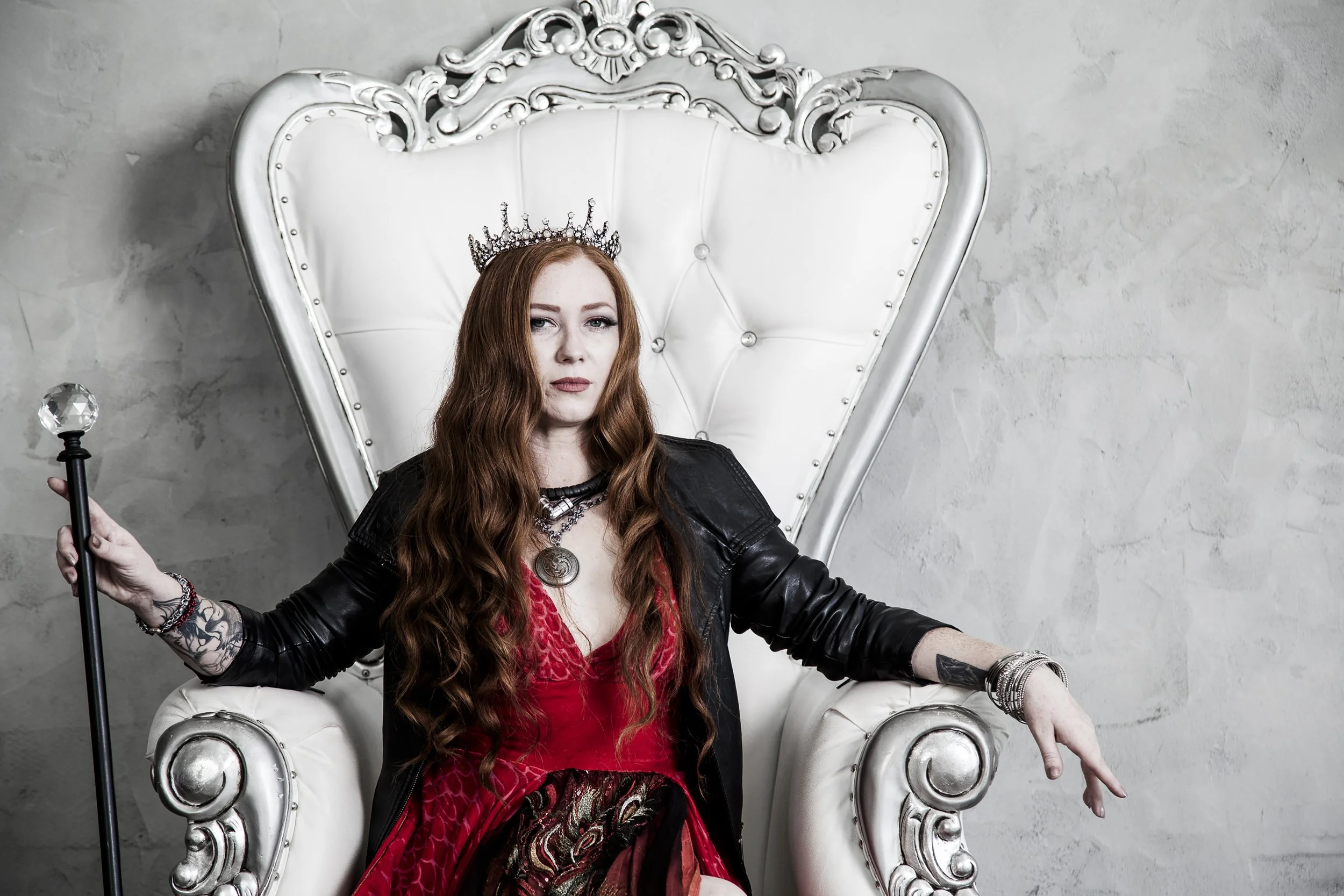 A red haired model sits in a white throne like chair in a studio. She is wearing a one of a kind red dress with peacock feather and a deeper red ombré hem. She is wearing a crown.