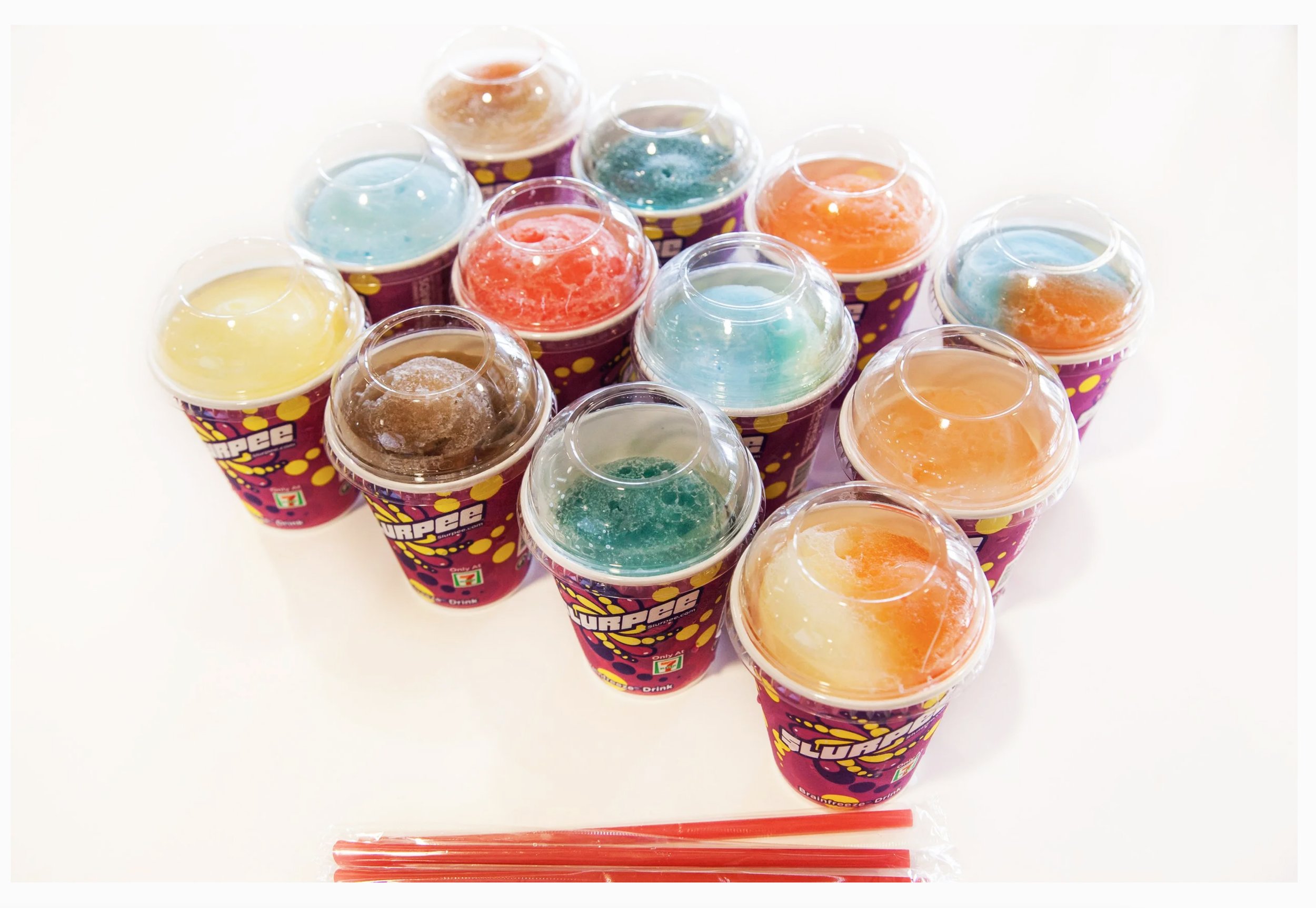 Display image of twelve (12) 7-Eleven Slurpees of a variety of colors and flavors. They are set up on a white table in a rectangle formation. There are red straws lying on the table in front of the cups. 