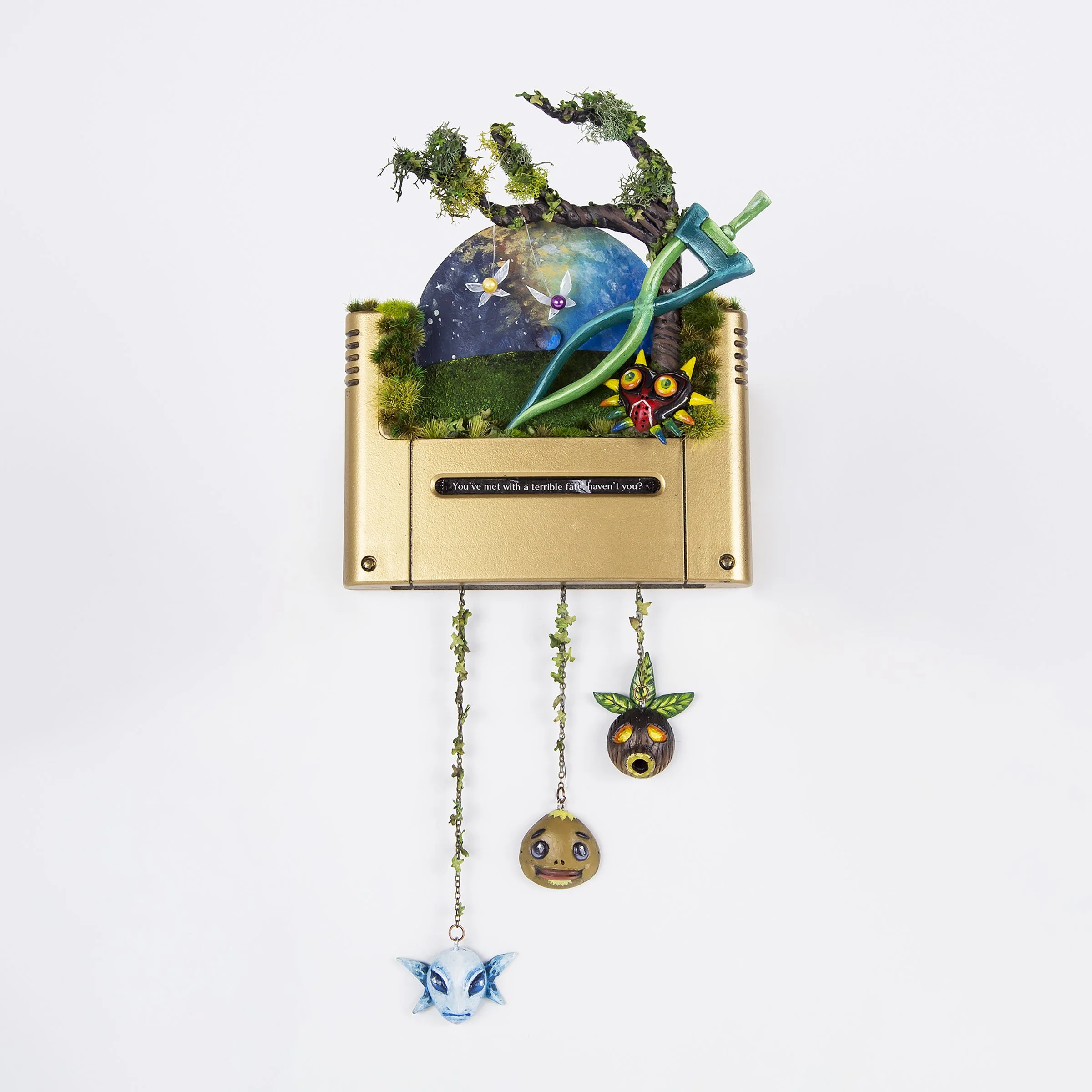 E-commerce and product image of a unique art piece based on a video game design. Piece includes a gold painted gaming cartridge, a rotating moonscape, tree branches and fabricated character pendants hanging from chains designed to look like vines.