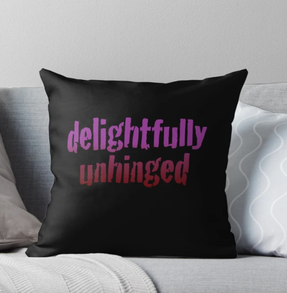 Black pillow with graphic wording on it reading Delightfully Unhinged in a pink to red ombré fade.