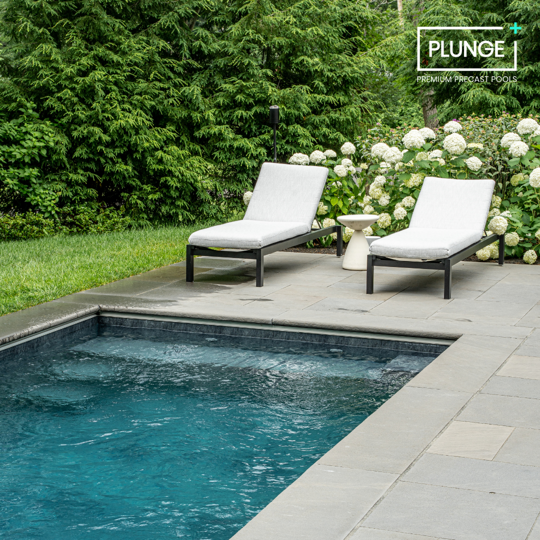 Two white cushioned lounge chairs next to a small white side table by a swimming pool, with lush green trees and white Hydrangea flowers in the background. The image promotes Plunge premium precast pools.